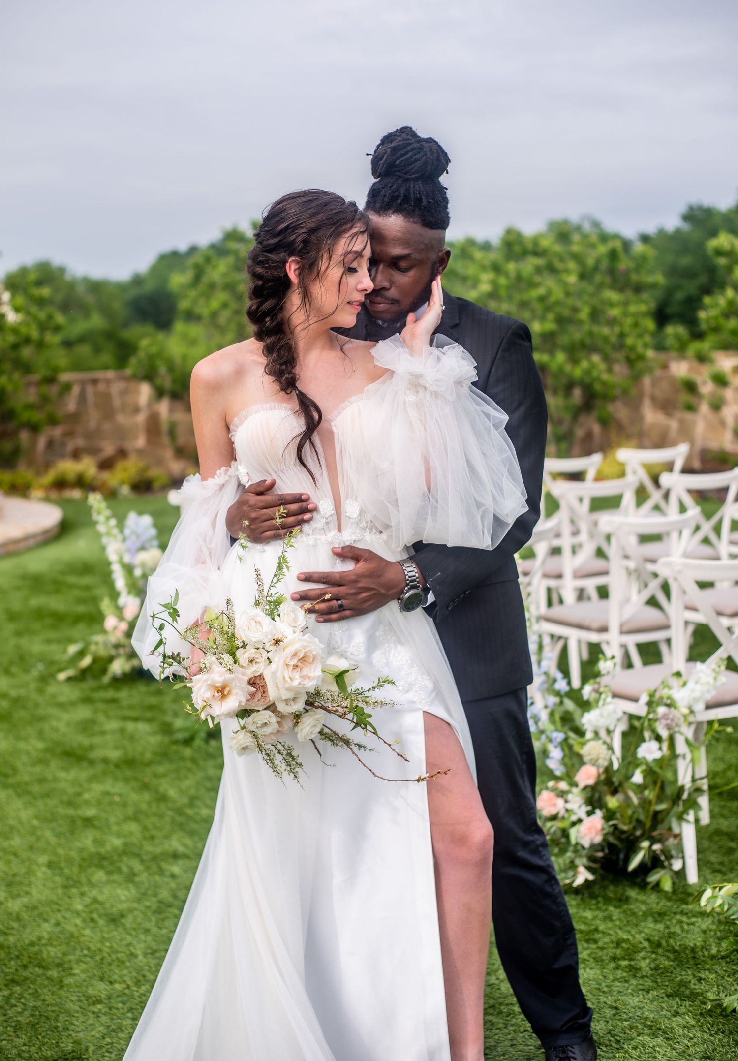 An interracial couple holds each other closely while the woman places her hand on his face. They are wearing wedding attire