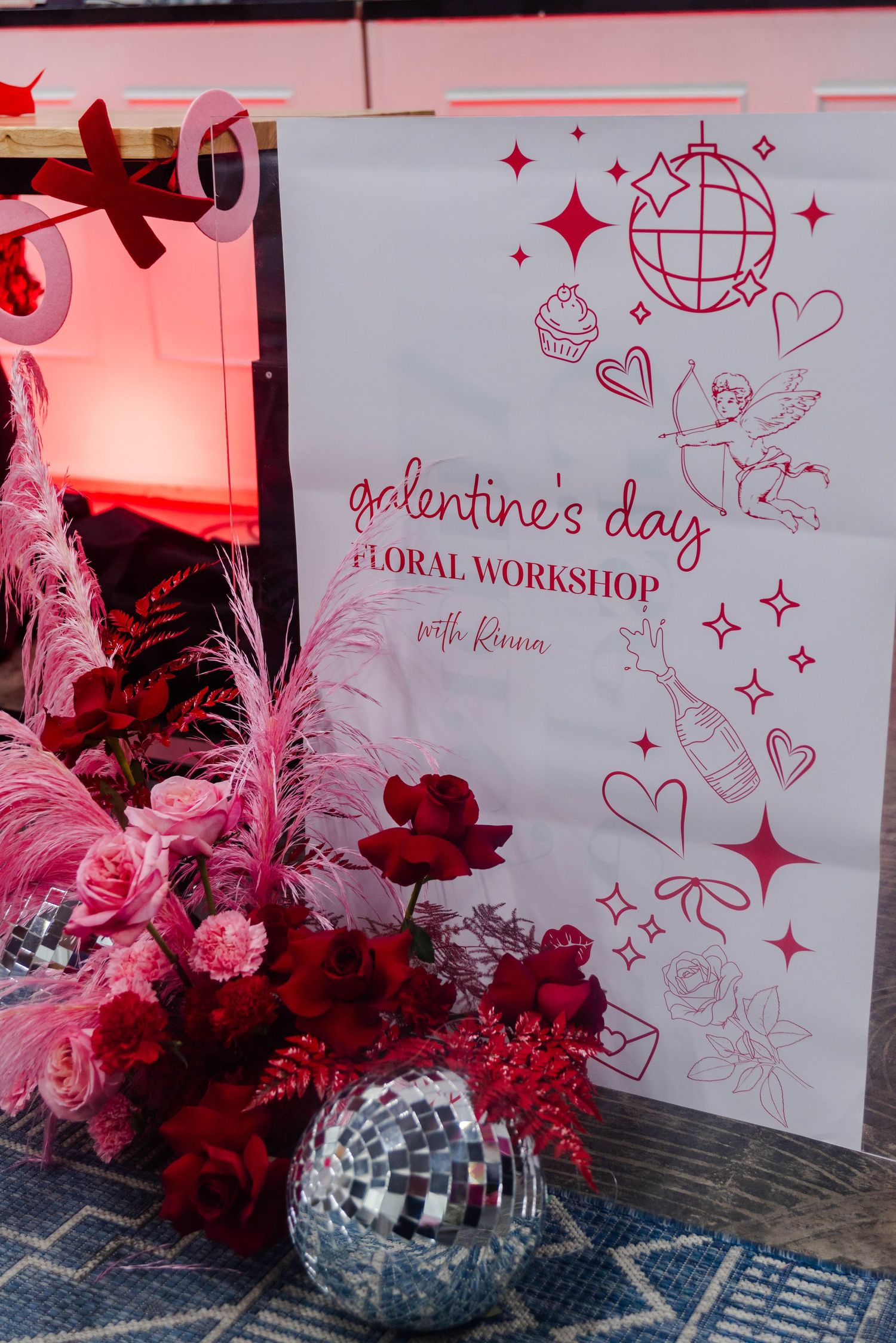 A Valentine's Day floral workshop poster with red decorations, pink feathers, and a disco ball displayed in a festive setup.