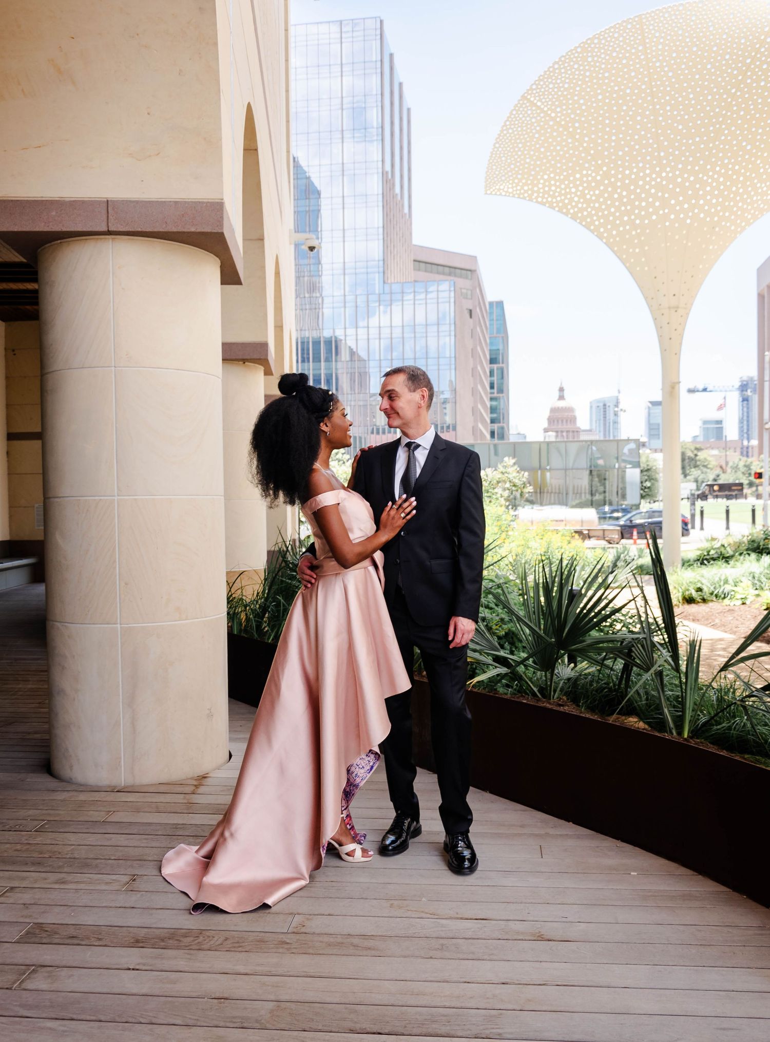 A couple in formal attire sharing a romantic moment on an outdoor terrace with a modern city skyline in the background.