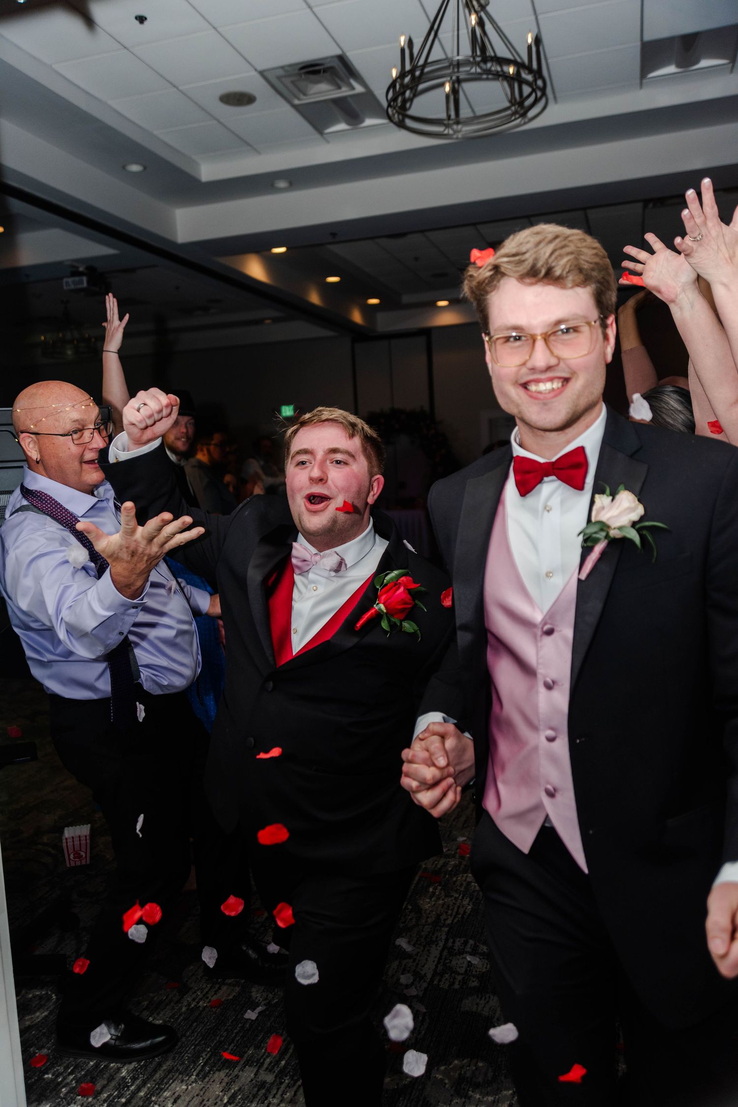 Two men hold each others hands and walk down the aisle as they exit their wedding, there are flower petals being thrown by guests