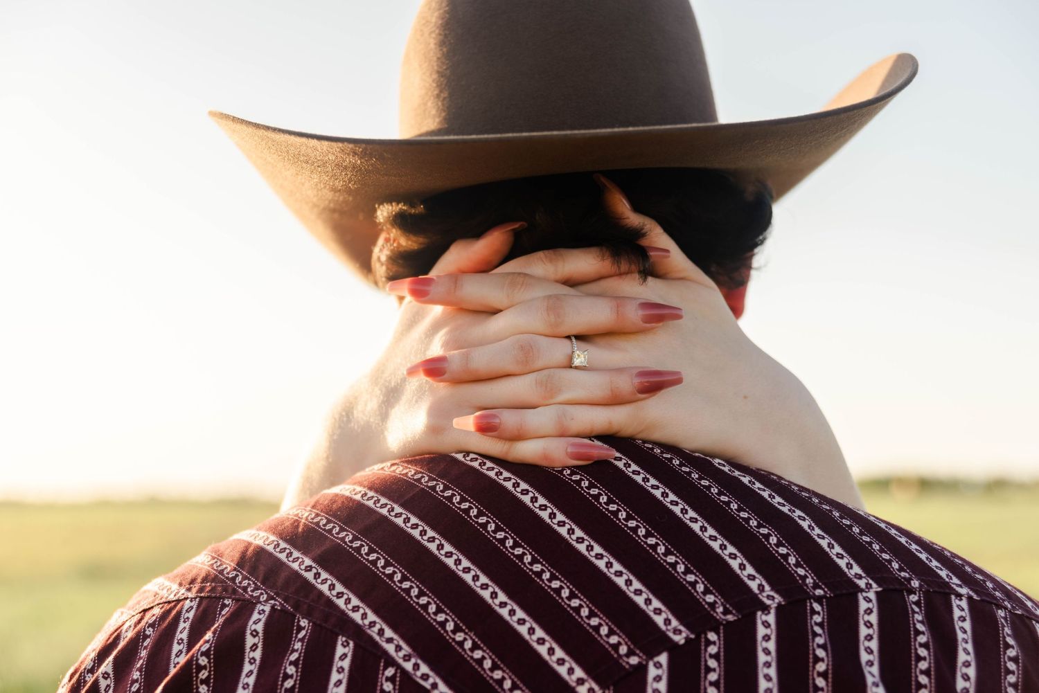womans hands wrapped around the back of the mans neck showing off her engagement ring. Man is wearing a western hat