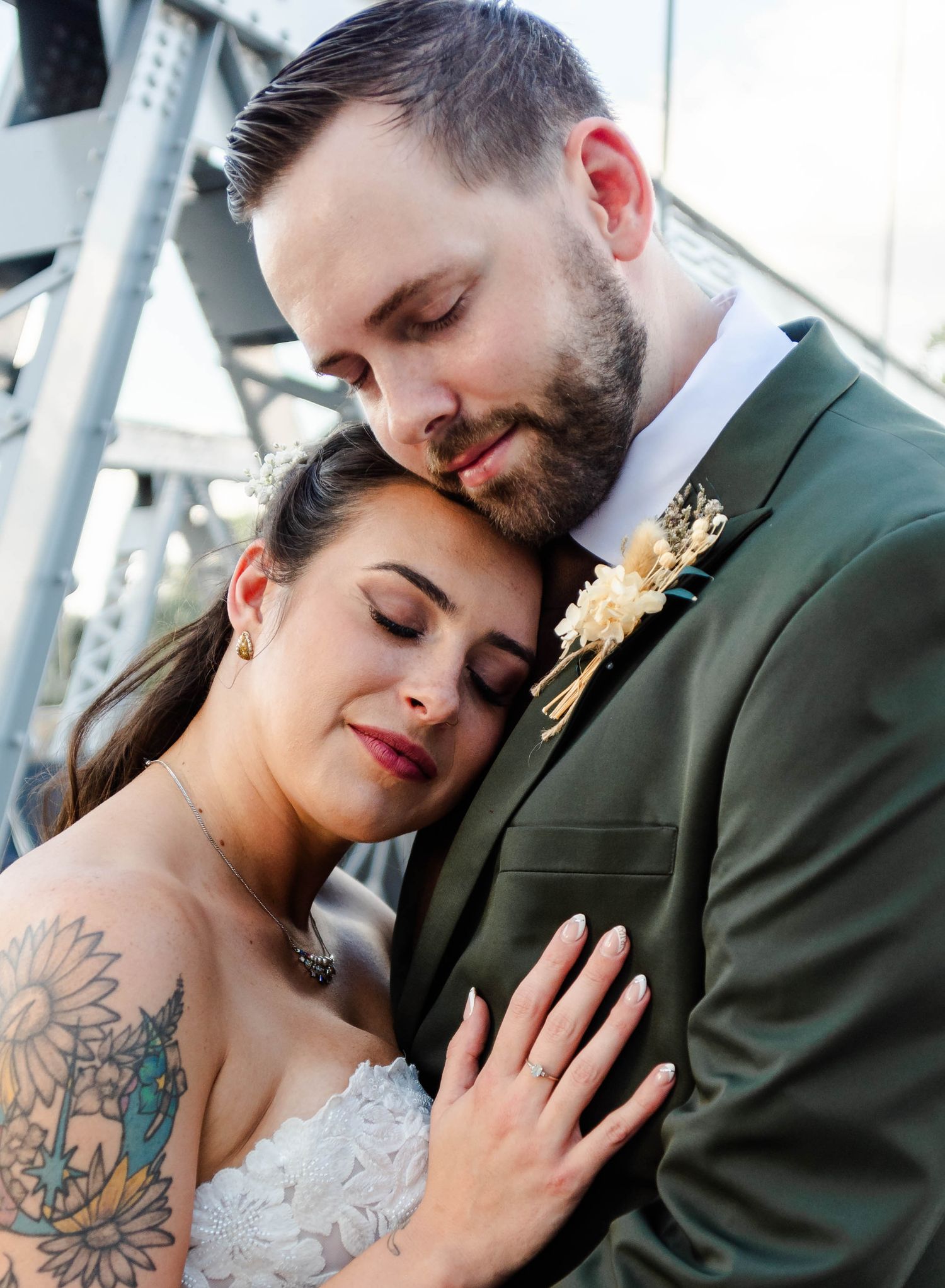A tender wedding moment captured against a metallic structure backdrop, with the bride displaying colorful arm tattoo art.
