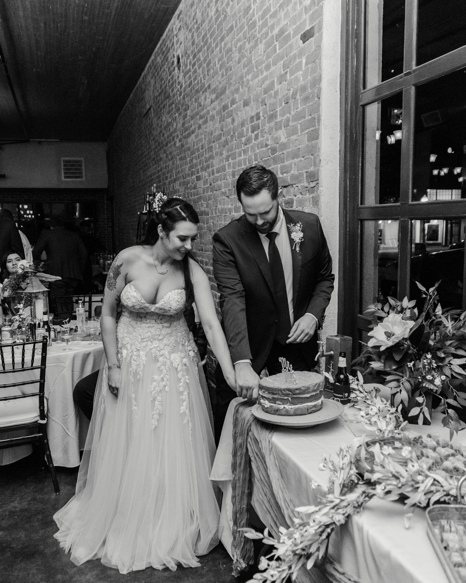 A bride and groom cut their wedding cake together at a reception with rustic brick wall and floral decor.