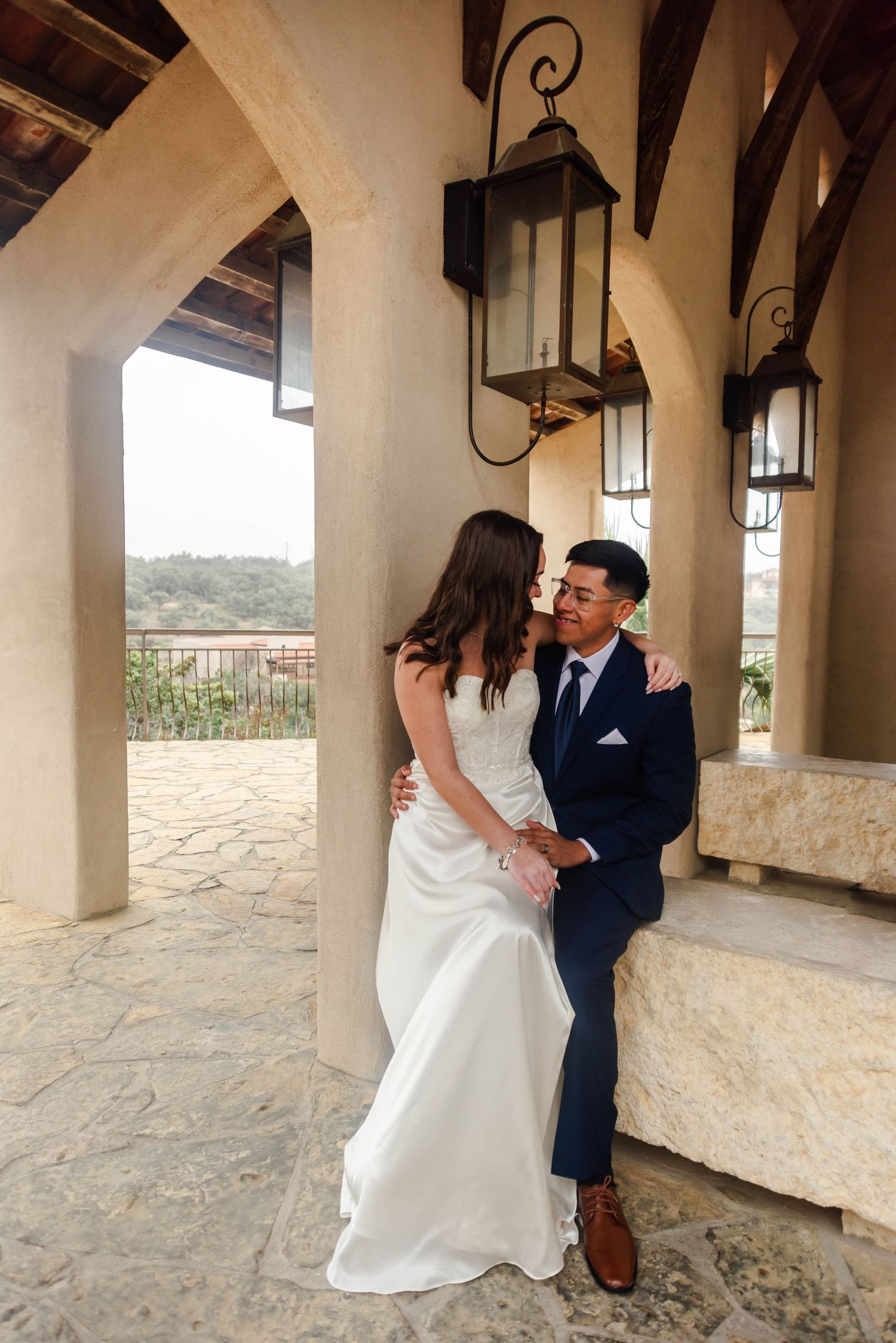 A romantic wedding couple embraces on a Mediterranean-style veranda with ornate lanterns and stone archways in the background.