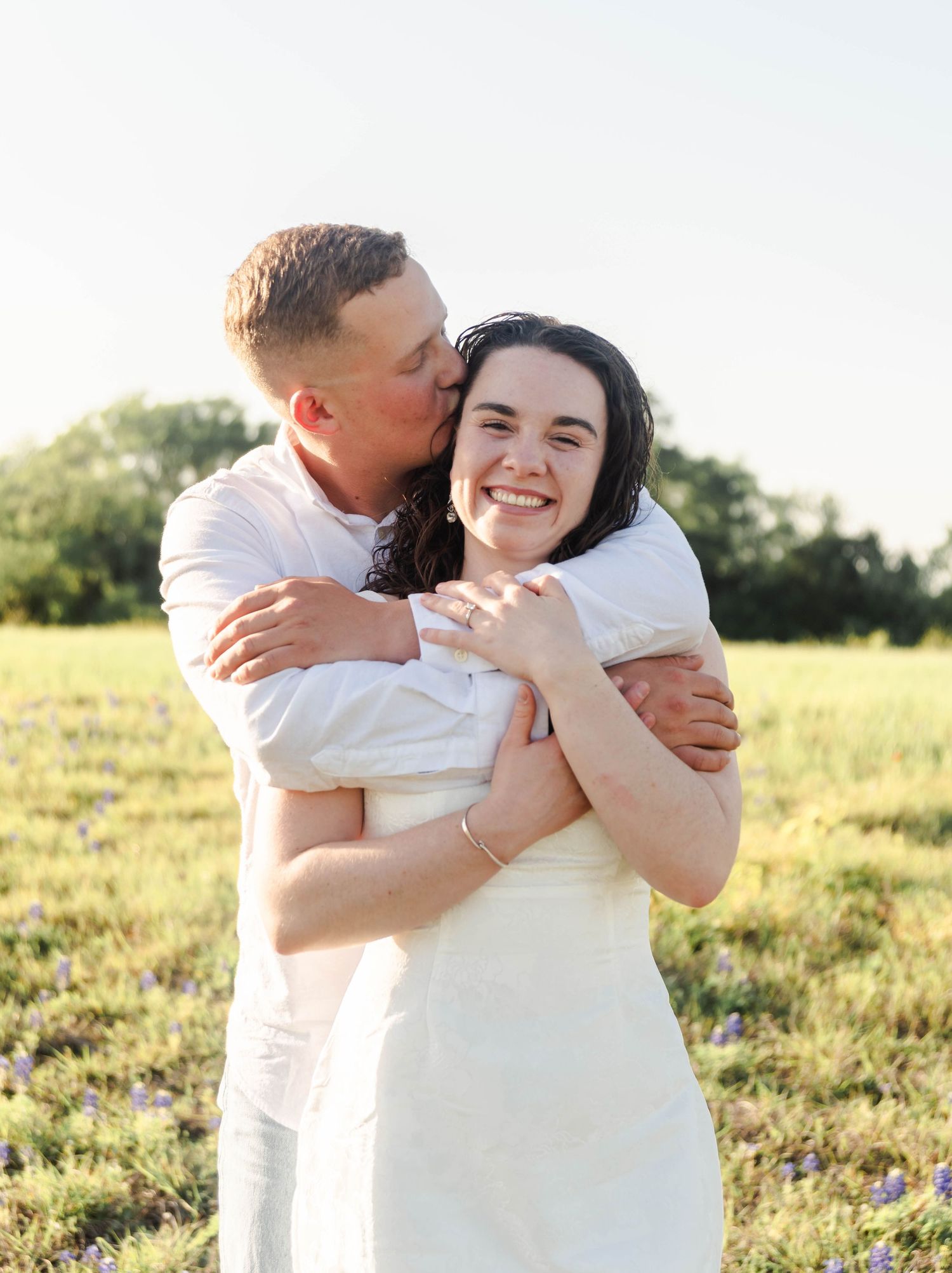 couple standing in an open field with arms wrapped around one another smiling.