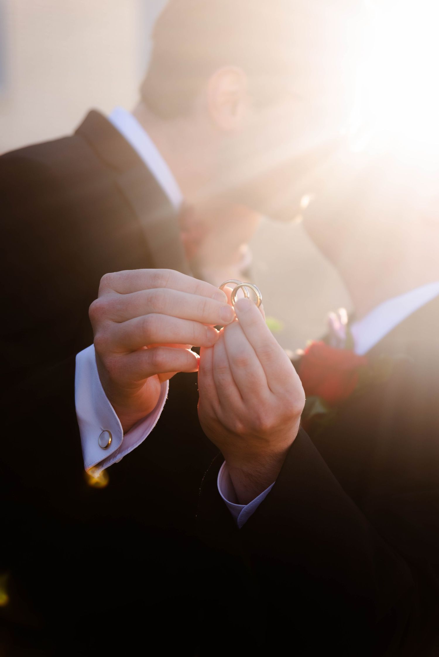 Two men hold their wedding rings together while giving each other a kiss. The sun has peaked through giving a starburst effect