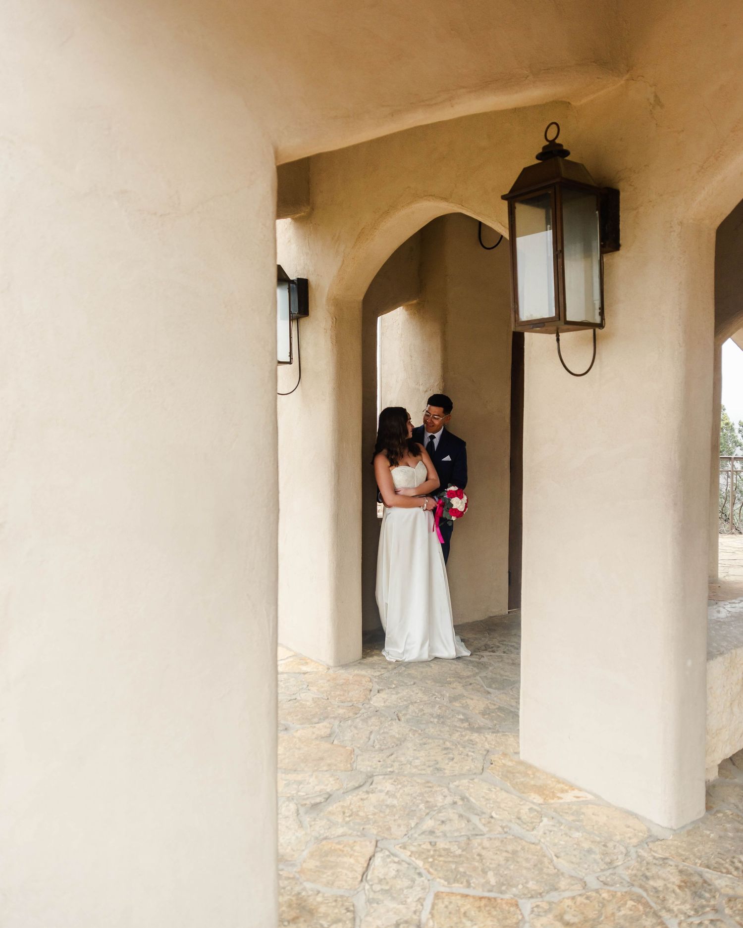 A couple stands together in an arched doorway of a Mediterranean-style building with stone flooring and hanging lanterns.