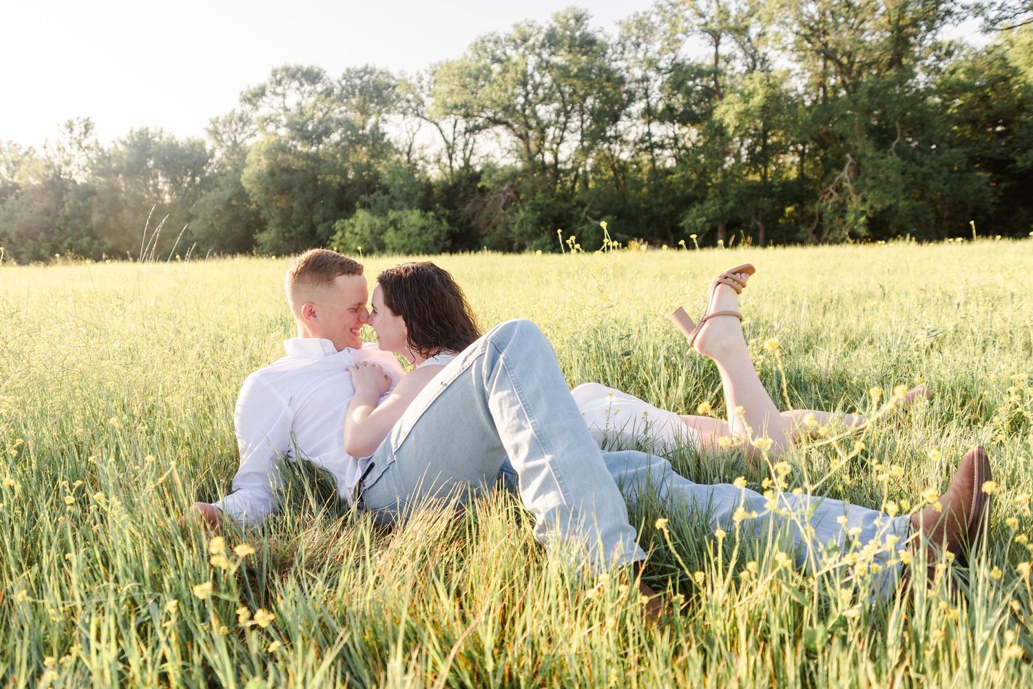 couple laying in a field with yellow flowers together smiling at each other, the woman has her leg kicked up.