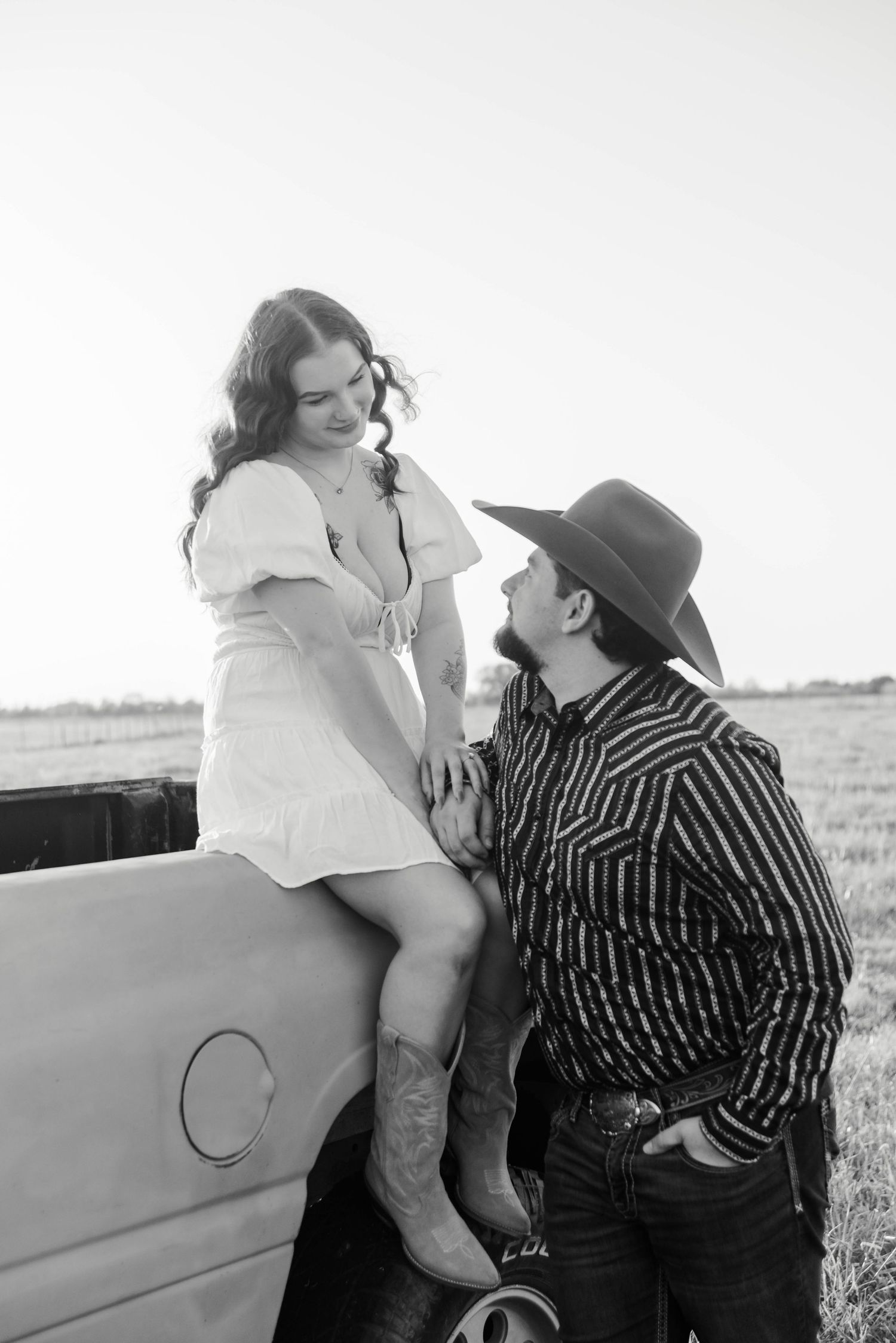 woman is sitting on the edge of a pickup truck bed as the man looks up at her lovingly