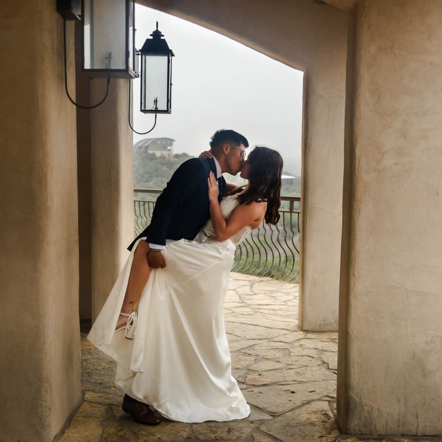 A couple sharing a romantic kiss in an archway on a stone balcony with a lantern mounted on the wall.