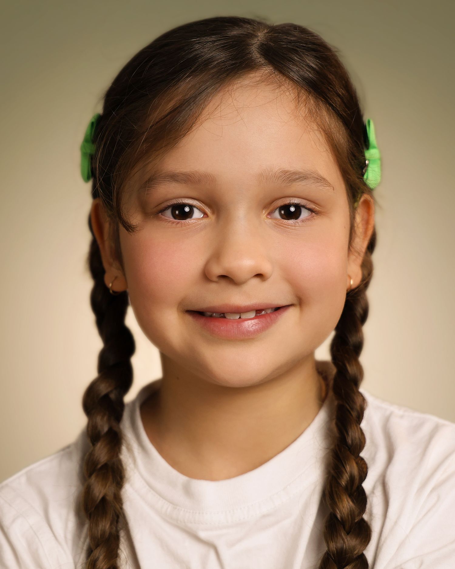Young girl with brown braids and green hair clips smiles warmly in a white shirt portrait photo.