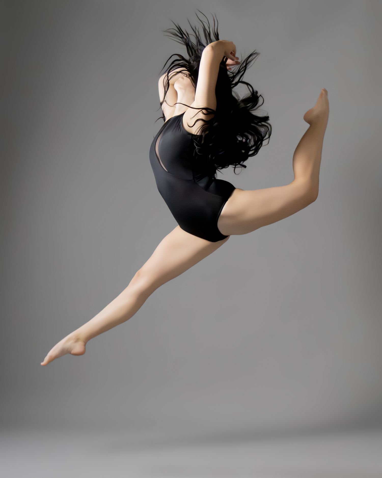 A dancer leaps gracefully in a black leotard, long dark hair flowing, against a gray studio background.