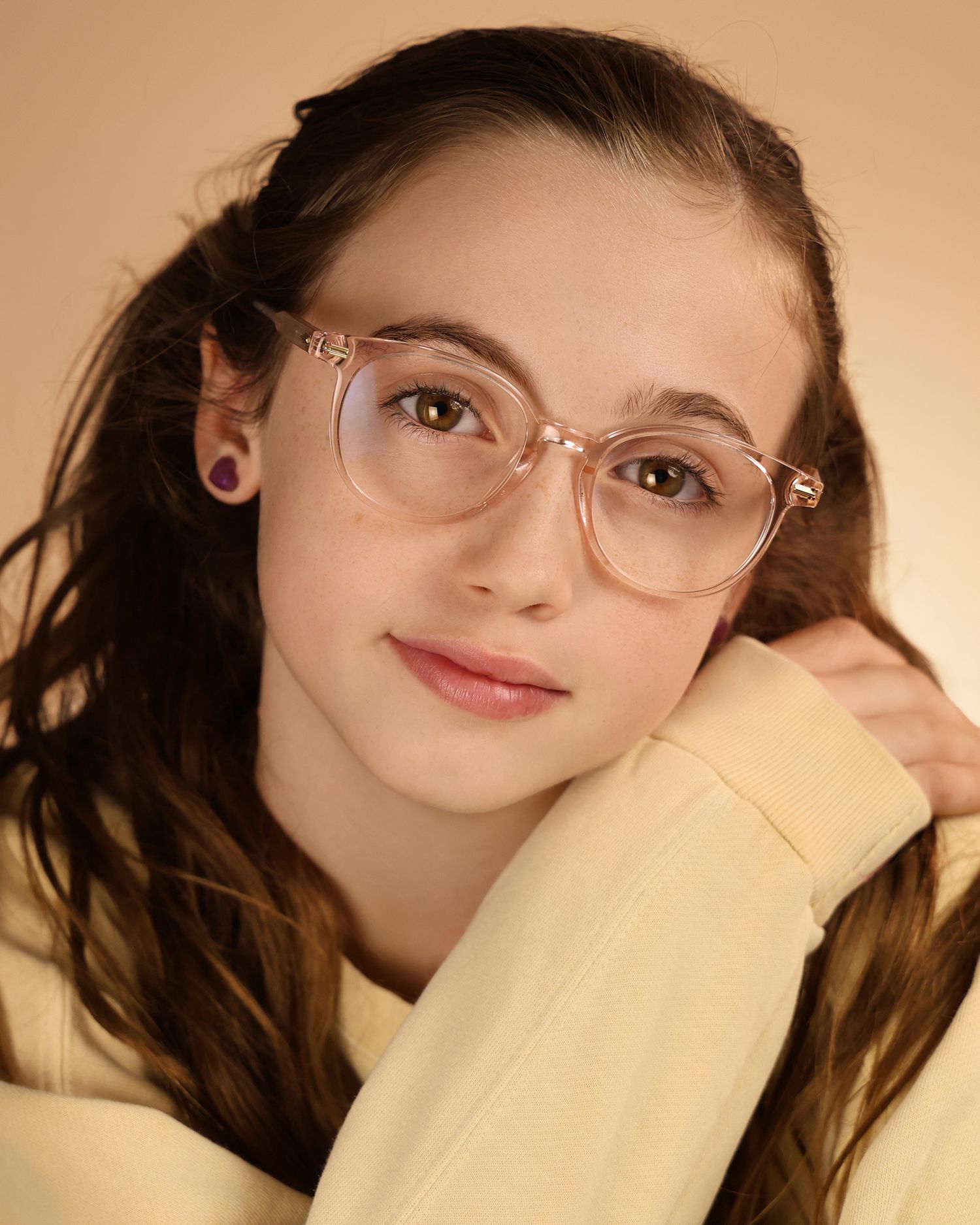 Young woman with long brown hair wearing clear pink glasses and a pale yellow hoodie poses against a beige background.