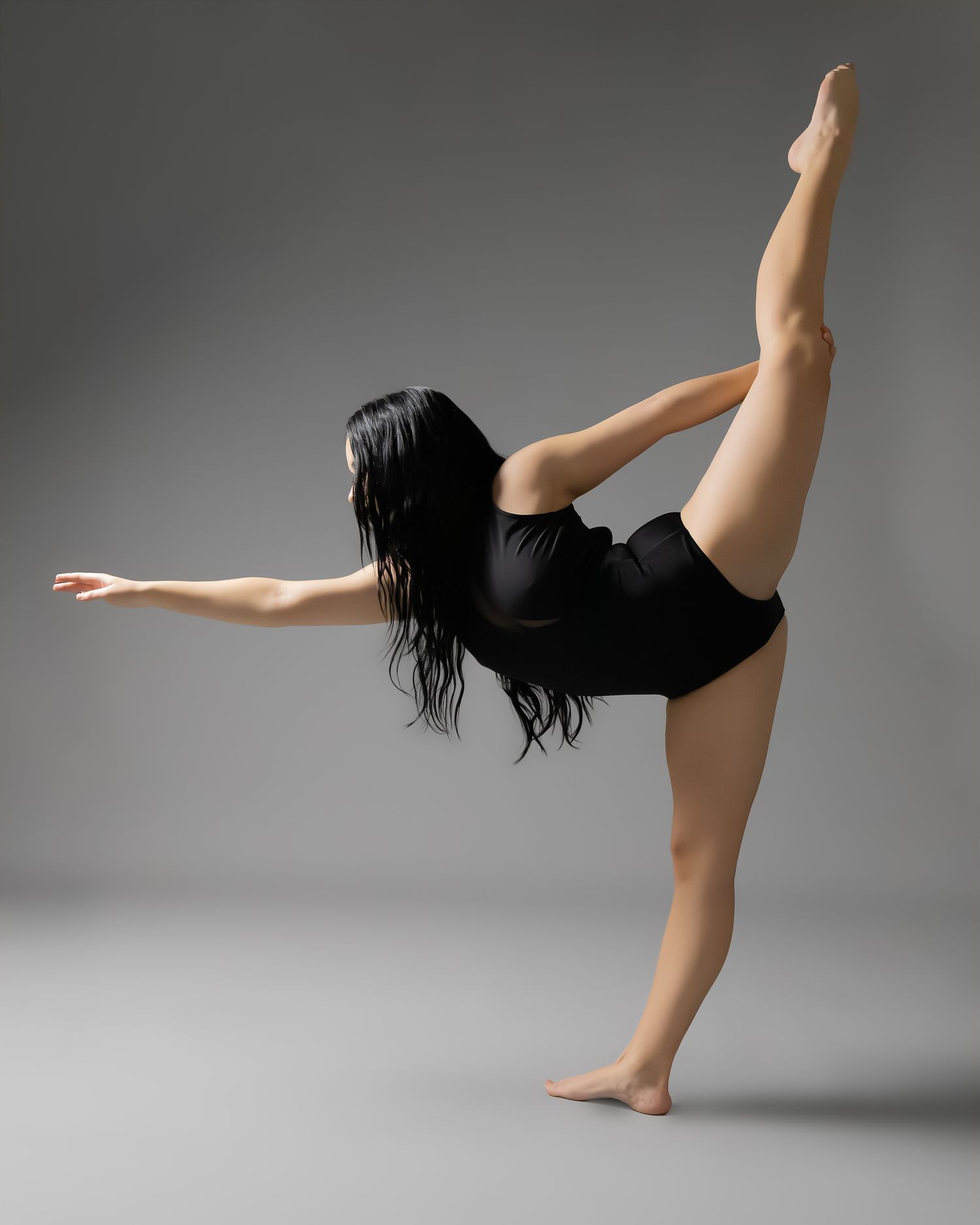 A flexible dancer in a black leotard balances on one foot while extending her leg high in a graceful studio pose.