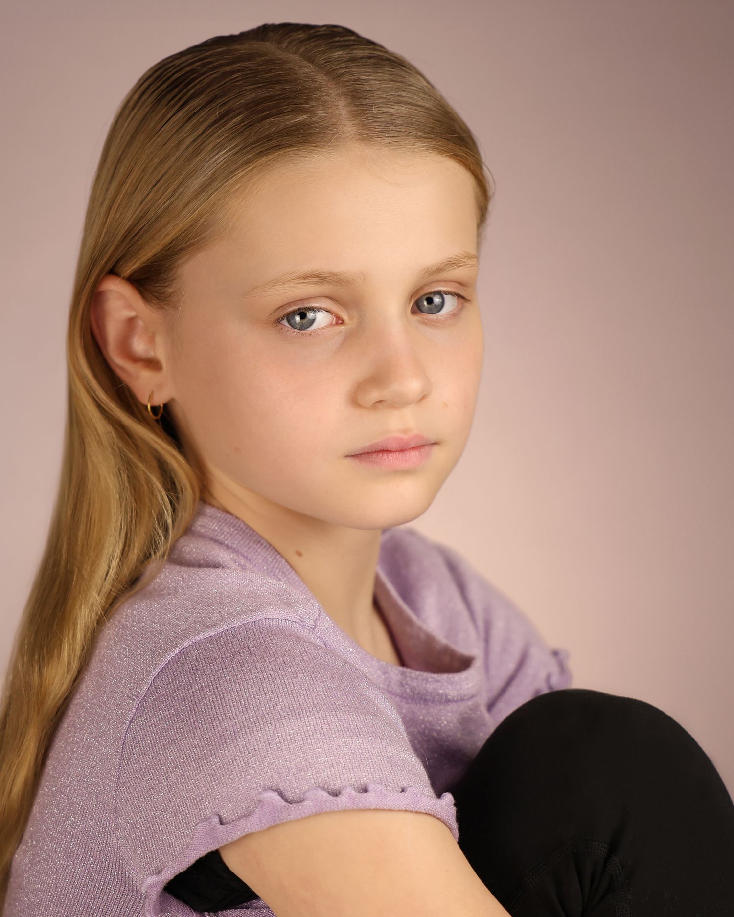 Young girl with blonde hair in lavender top poses for a portrait photo against a soft pink background.
