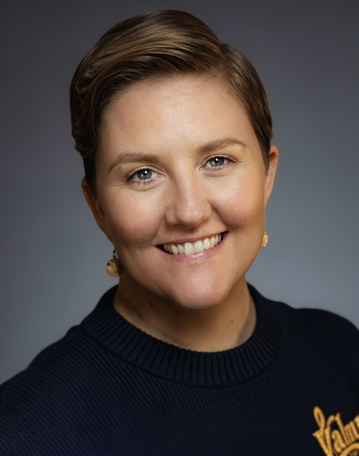 Professional headshot of a smiling woman with short brown hair wearing a navy blue sweater with gold logo.