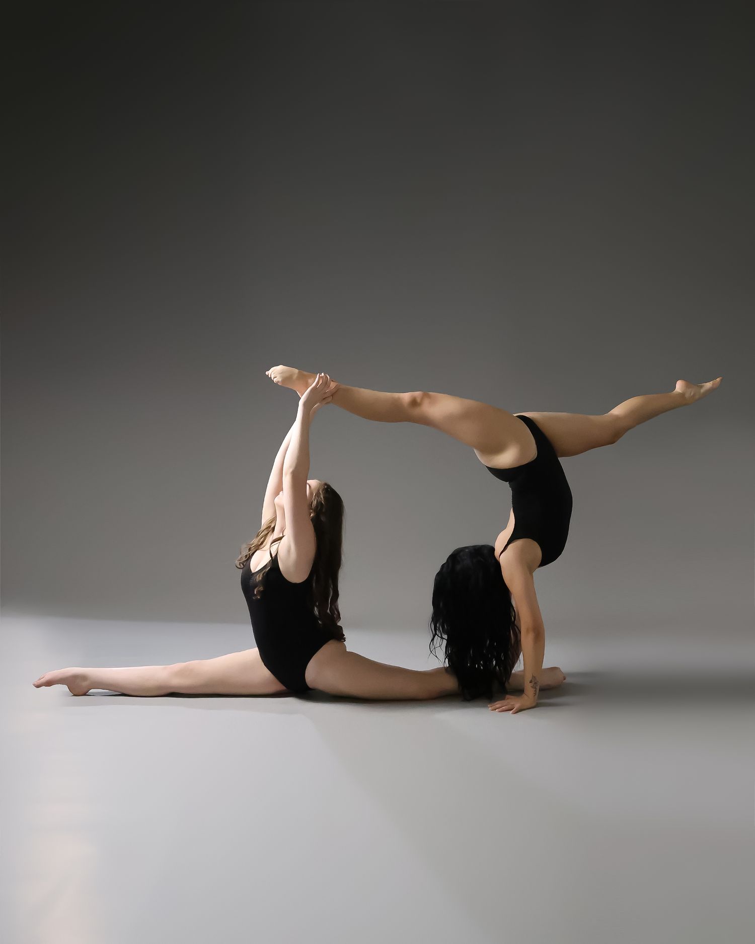 Two female dancers in black leotards perform an acrobatic partner pose on a gray studio floor.