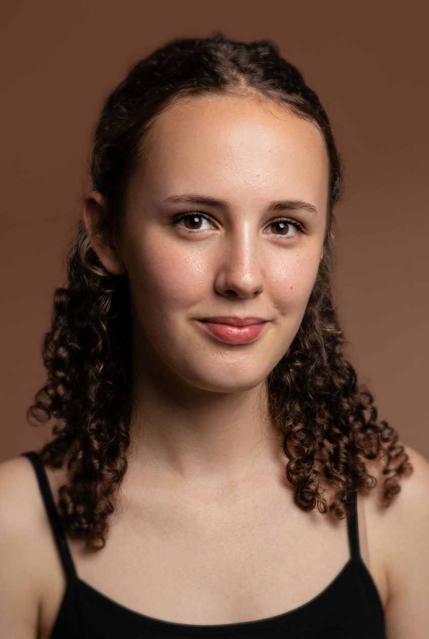Young woman with curly brown hair and a black top poses for a studio portrait against a brown background.