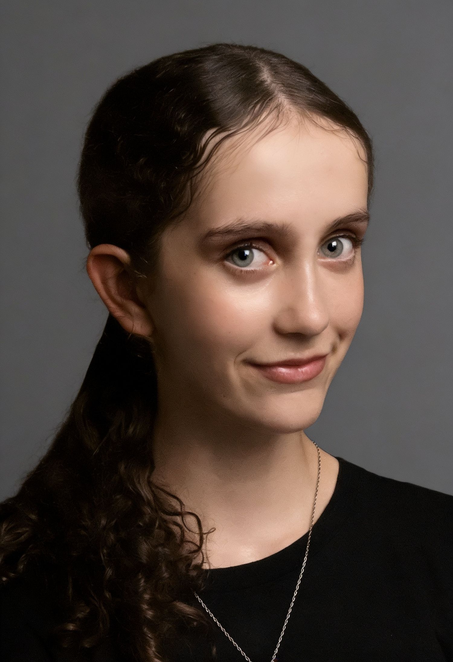 Young woman with curly dark hair in ponytail, wearing black top and silver necklace, smiling against gray background.