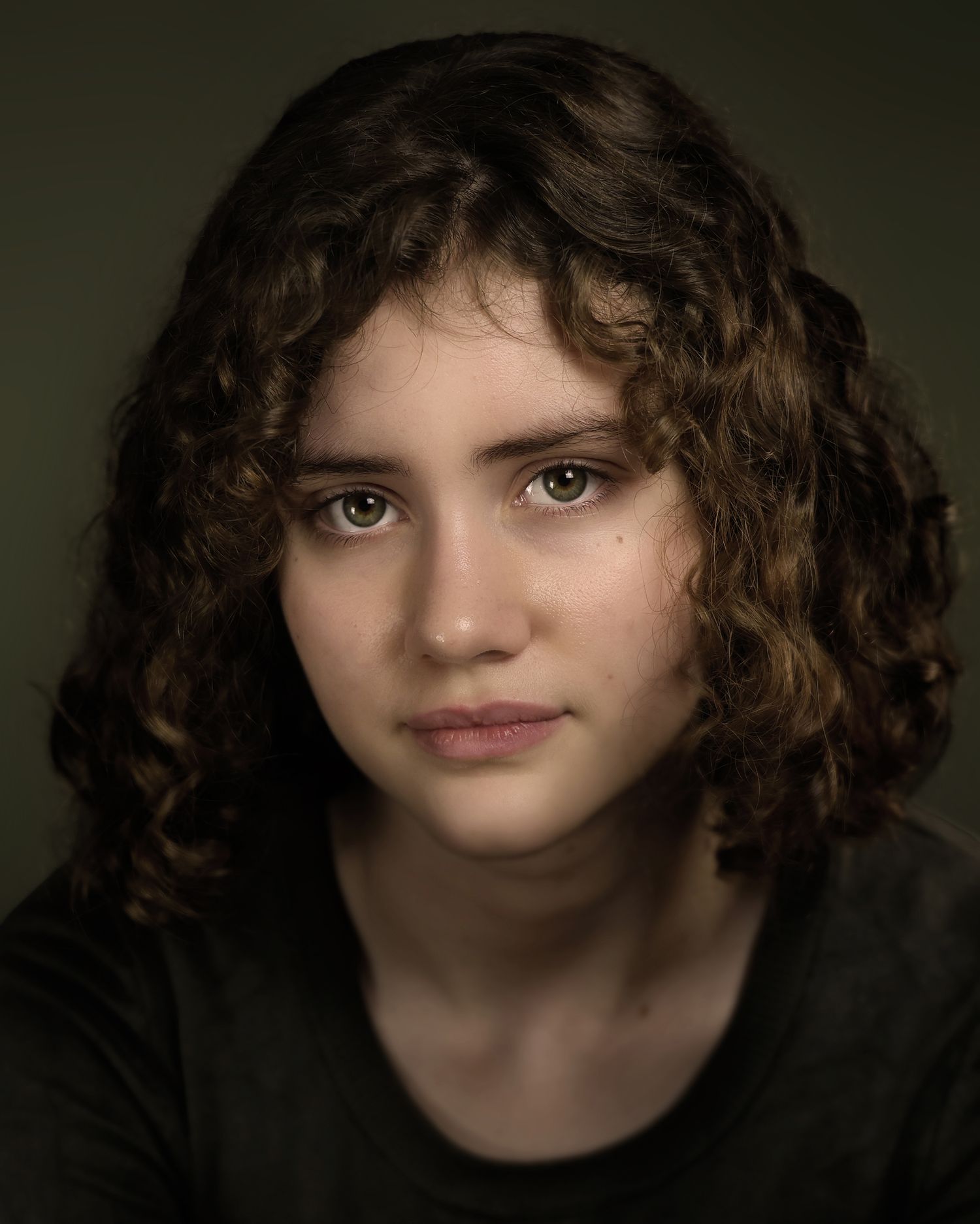 Young woman with curly brown hair and green eyes poses for a professional portrait against a dark background.