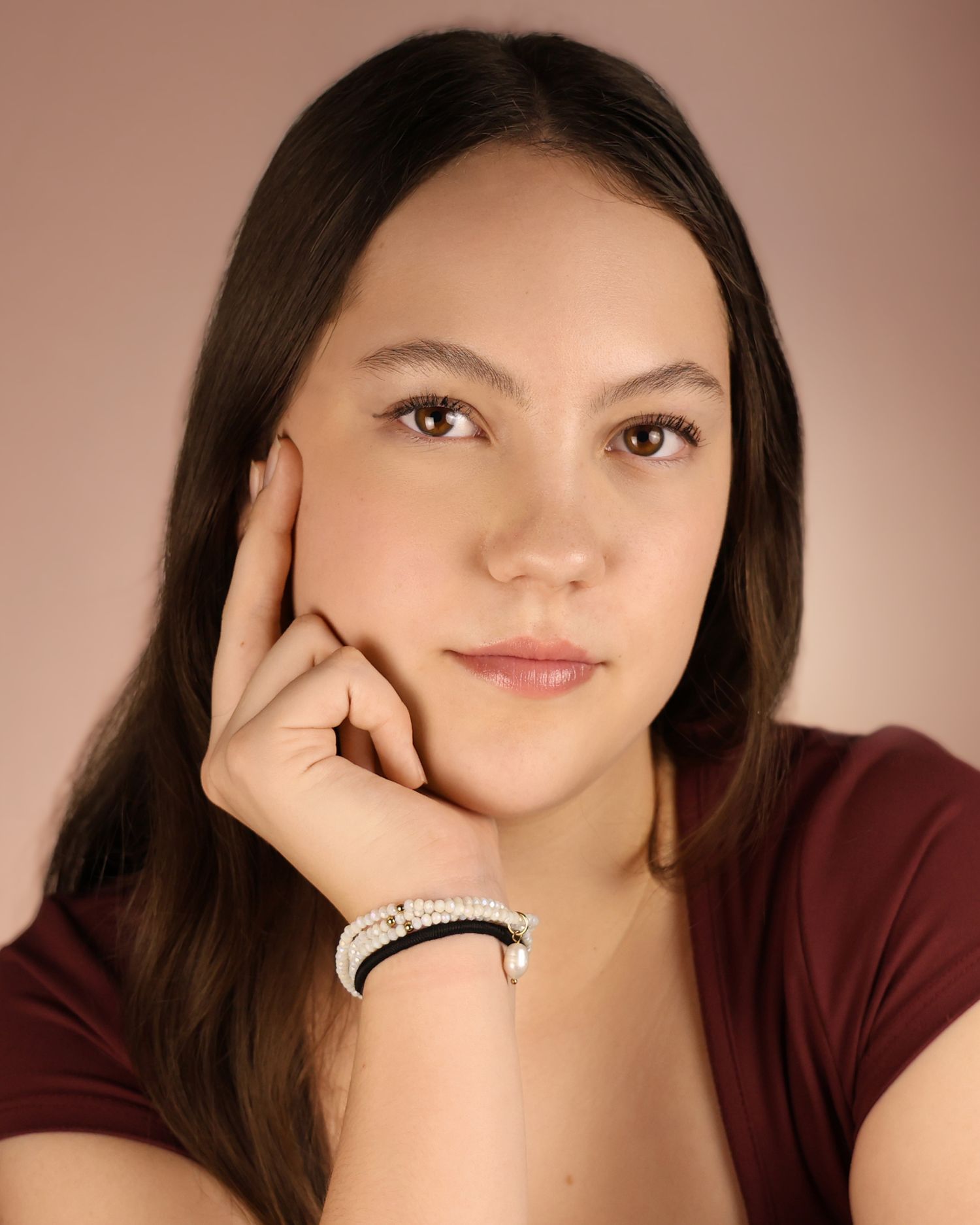Young woman with long dark hair resting chin on hand, wearing a maroon top and beaded bracelets, studio portrait.