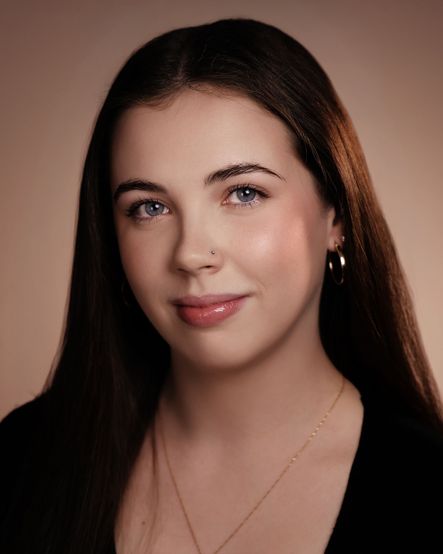 Young woman with blue eyes and long brown hair wearing a black top and gold jewelry against a neutral background.