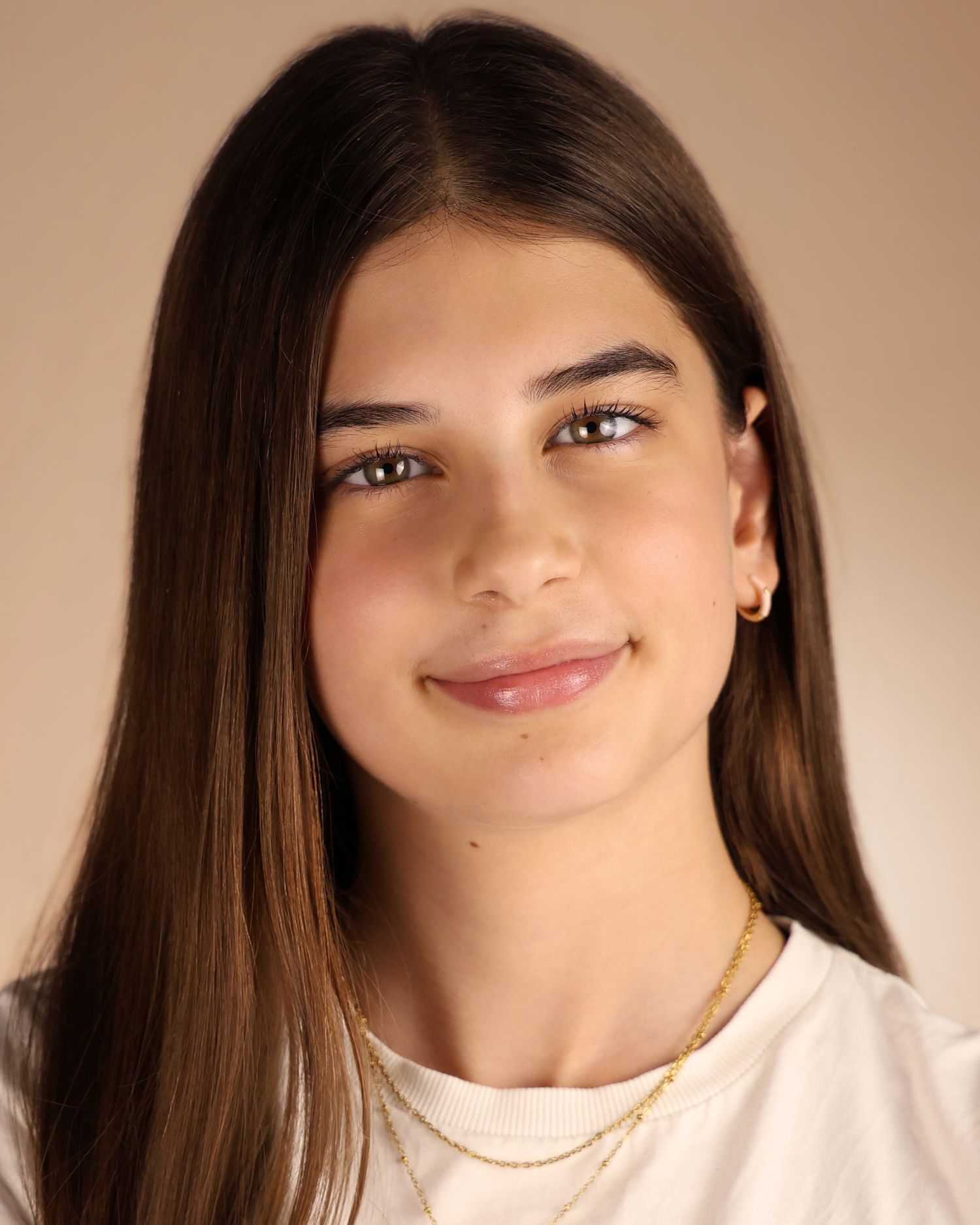 Young woman with long brown hair, gold necklace, and small earrings smiling softly against a neutral background.