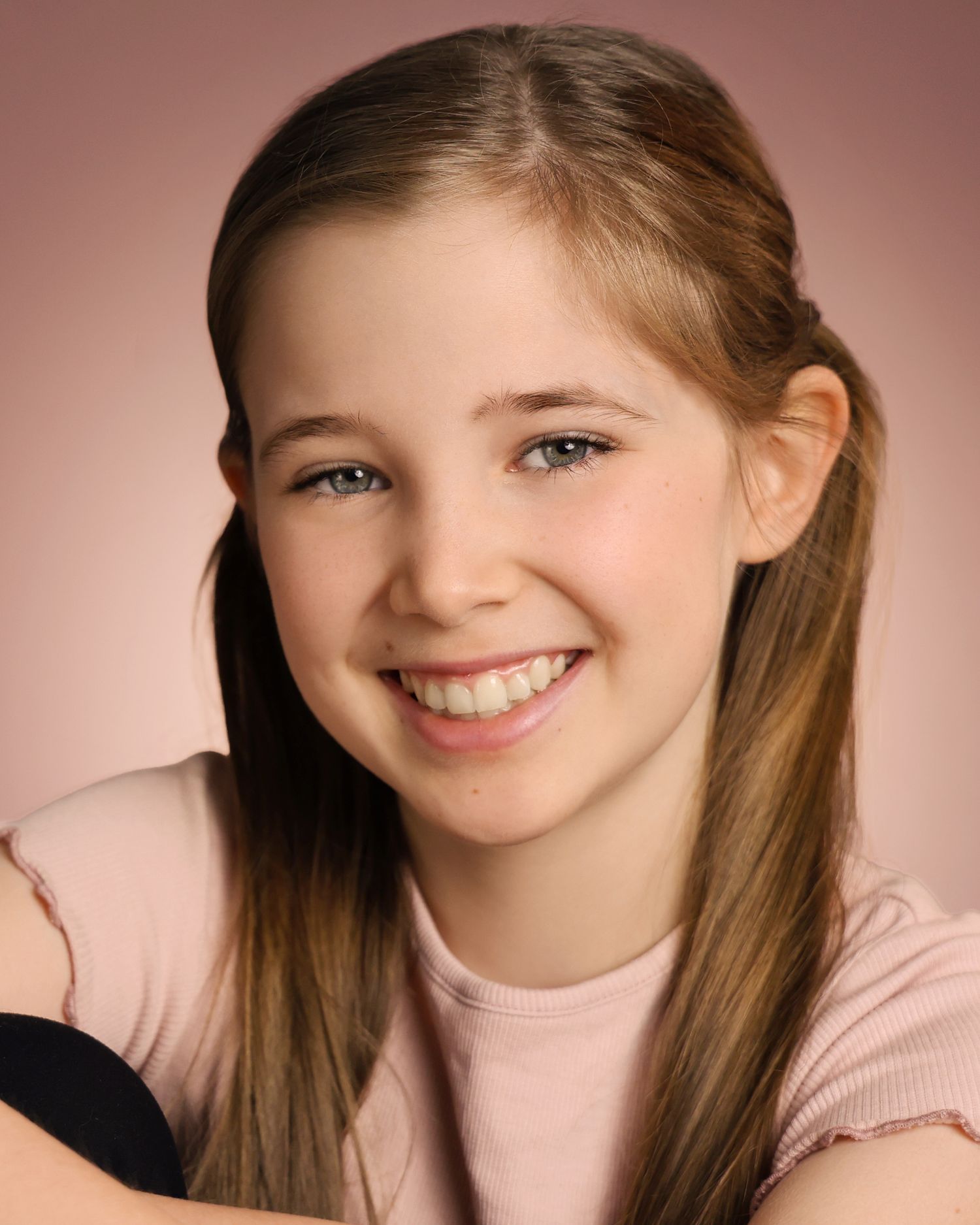 Smiling young girl with long brown hair wearing a pink top against a soft pink background.