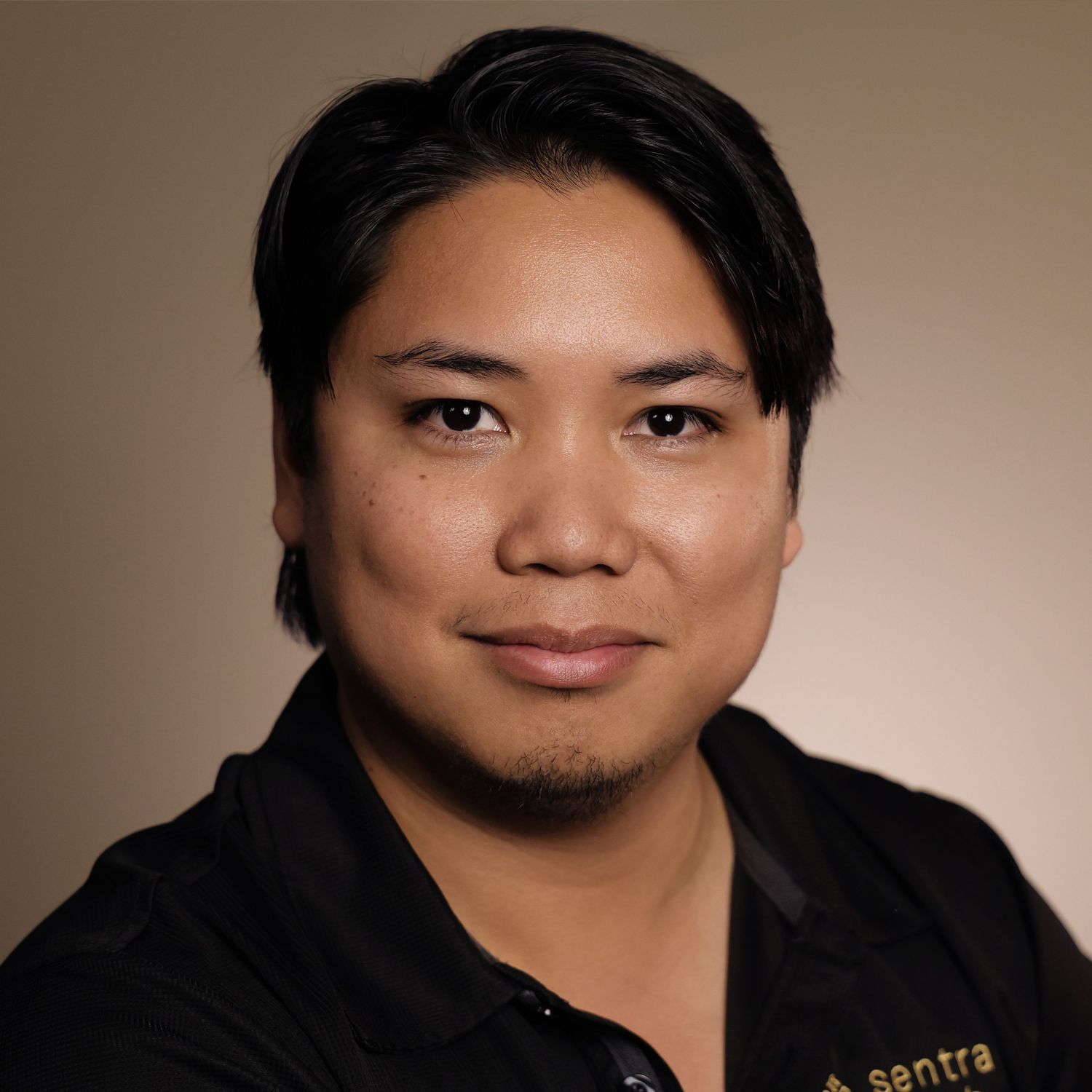 Professional headshot of a young Asian man with short dark hair and goatee wearing a black Sentra polo shirt.