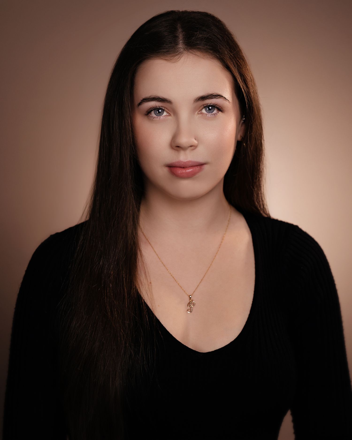 Young woman with long dark hair and blue eyes wearing a black top and delicate necklace against a warm background.