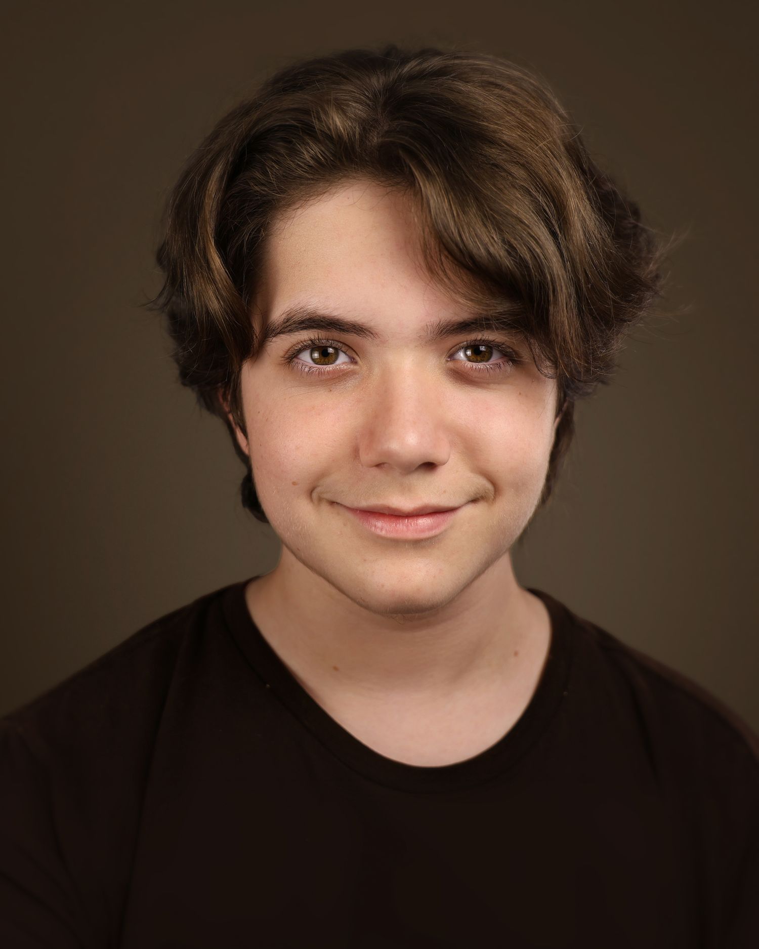 Young person with medium-length brown hair smiling warmly, wearing a black shirt against a dark studio background.