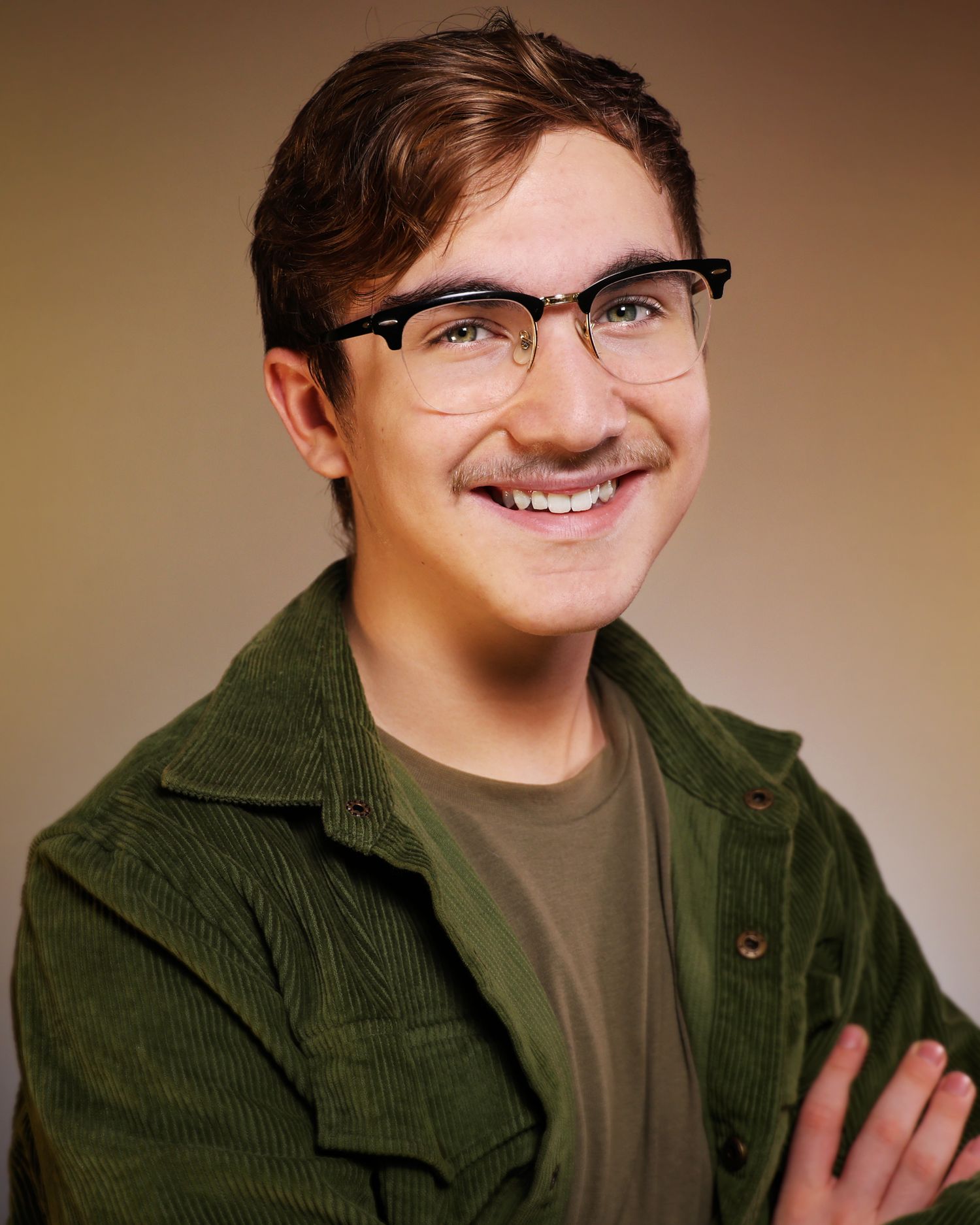 Young man with glasses and green corduroy jacket smiling confidently with arms crossed in a professional headshot.