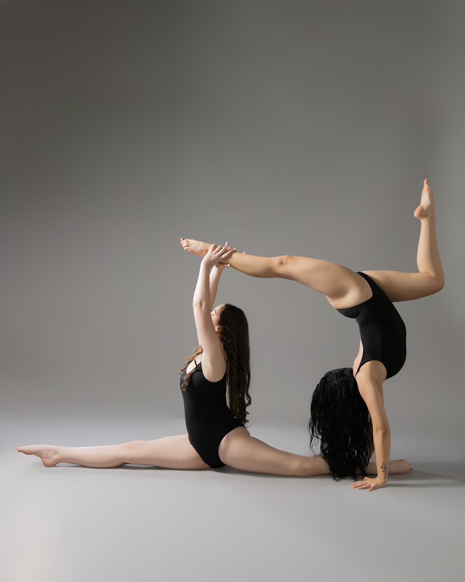 Two female dancers in black leotards perform an acrobatic pose together on a gray studio background.