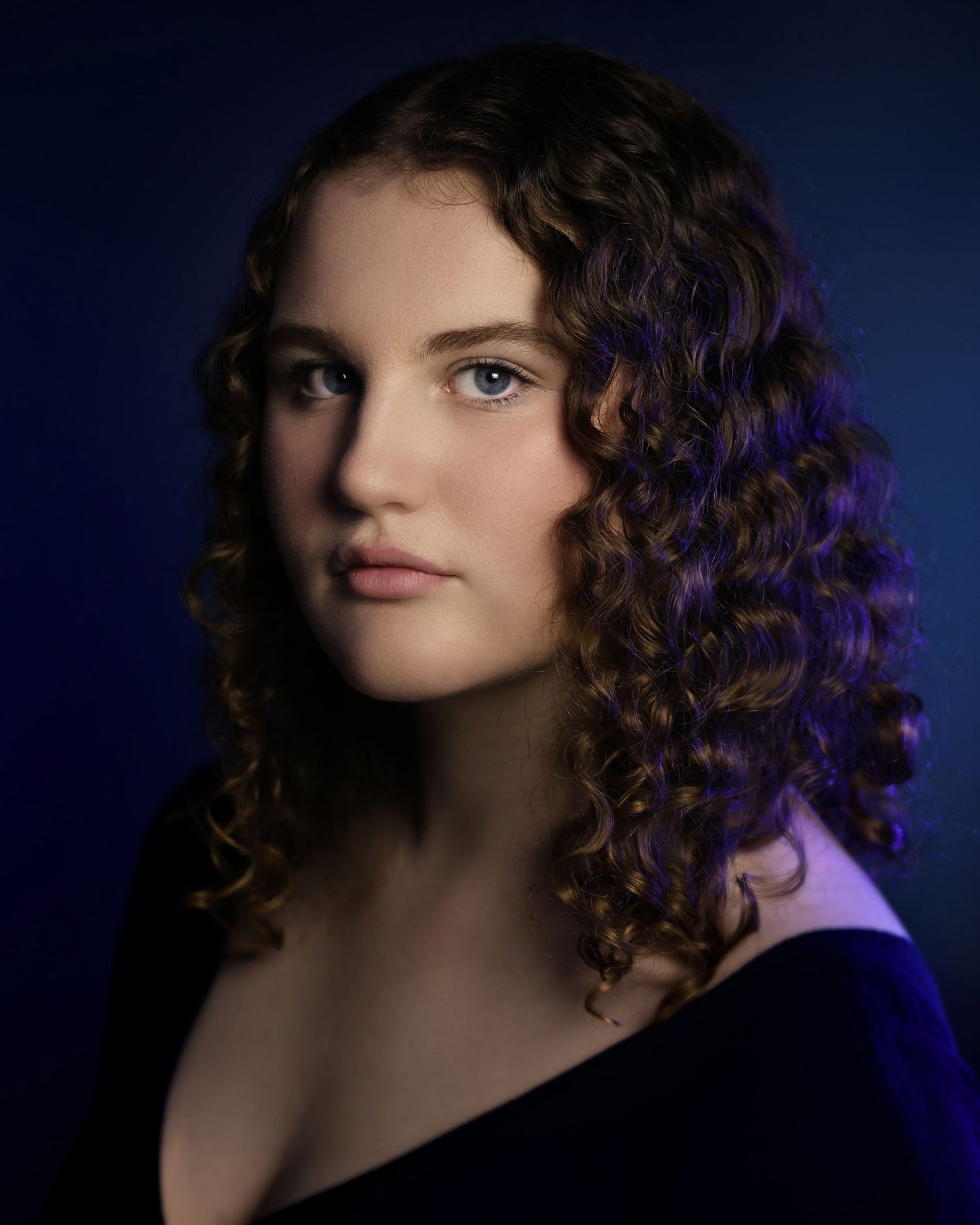 Young woman with curly brown hair and blue eyes poses against a dark blue background in a portrait photo.