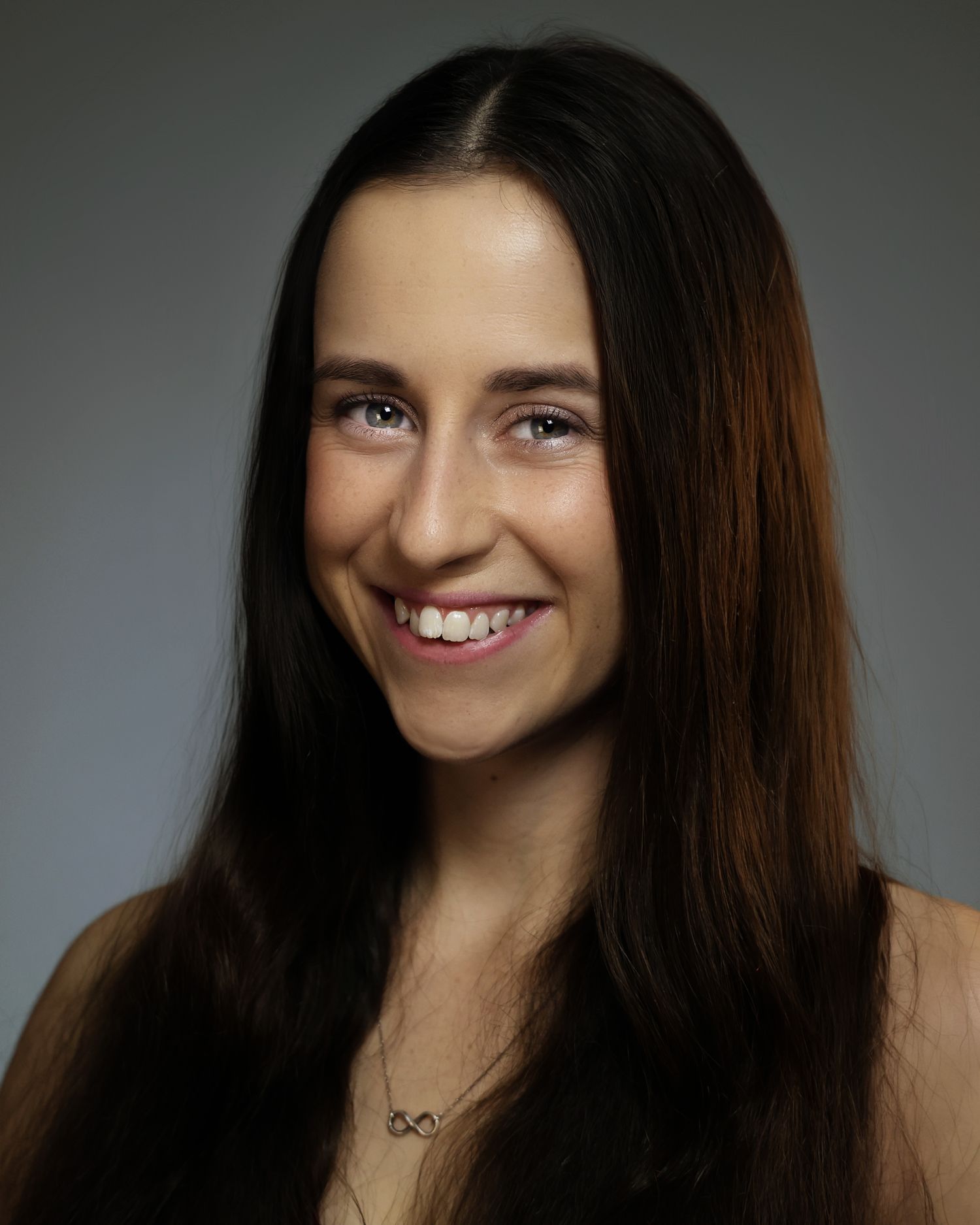 Young woman with long dark hair smiling warmly, wearing a delicate necklace, professional headshot photo.