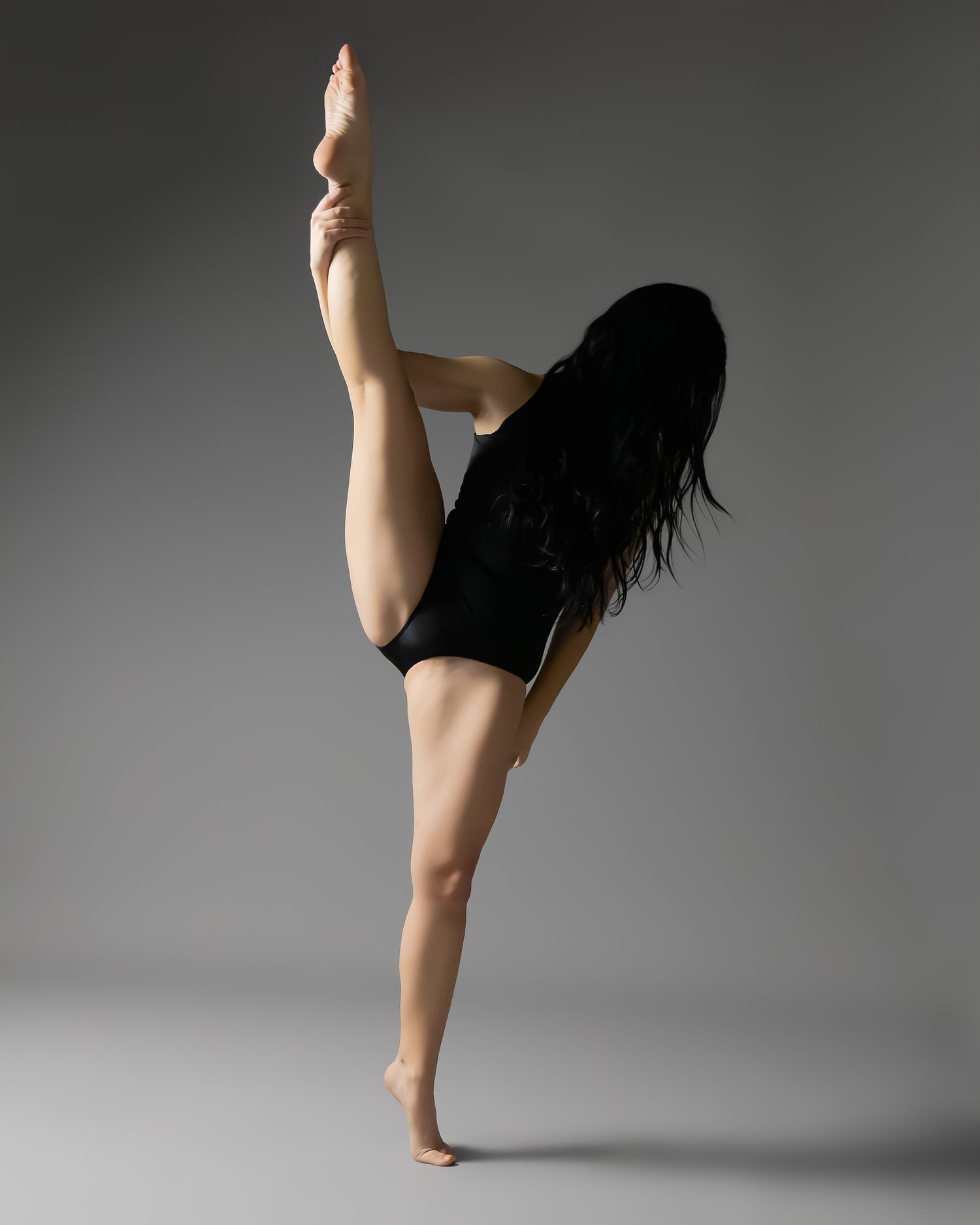 A flexible dancer in black leotard performs a standing split, raising one leg vertically while balancing on one foot.
