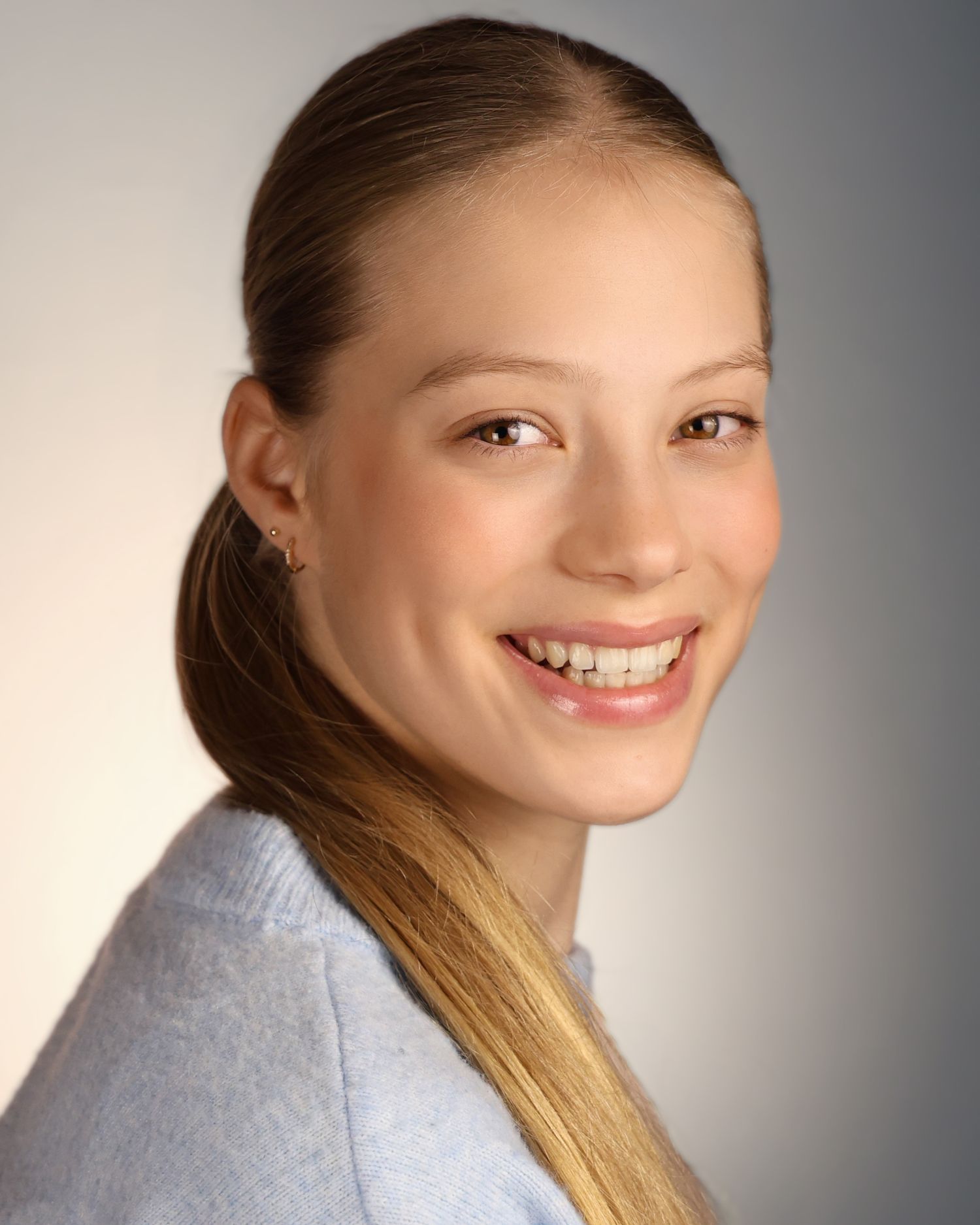 Smiling young woman with long blonde hair in a ponytail, wearing a light blue sweater, posing for a headshot photo.