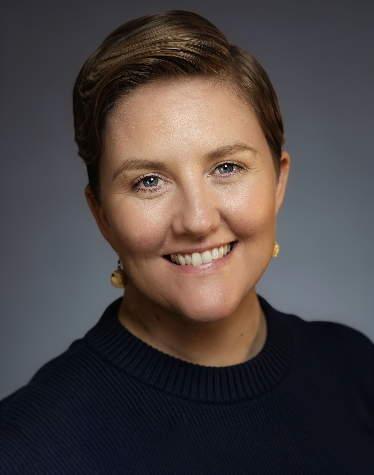 Professional headshot of a smiling woman with short brown hair wearing a navy sweater and gold earrings.