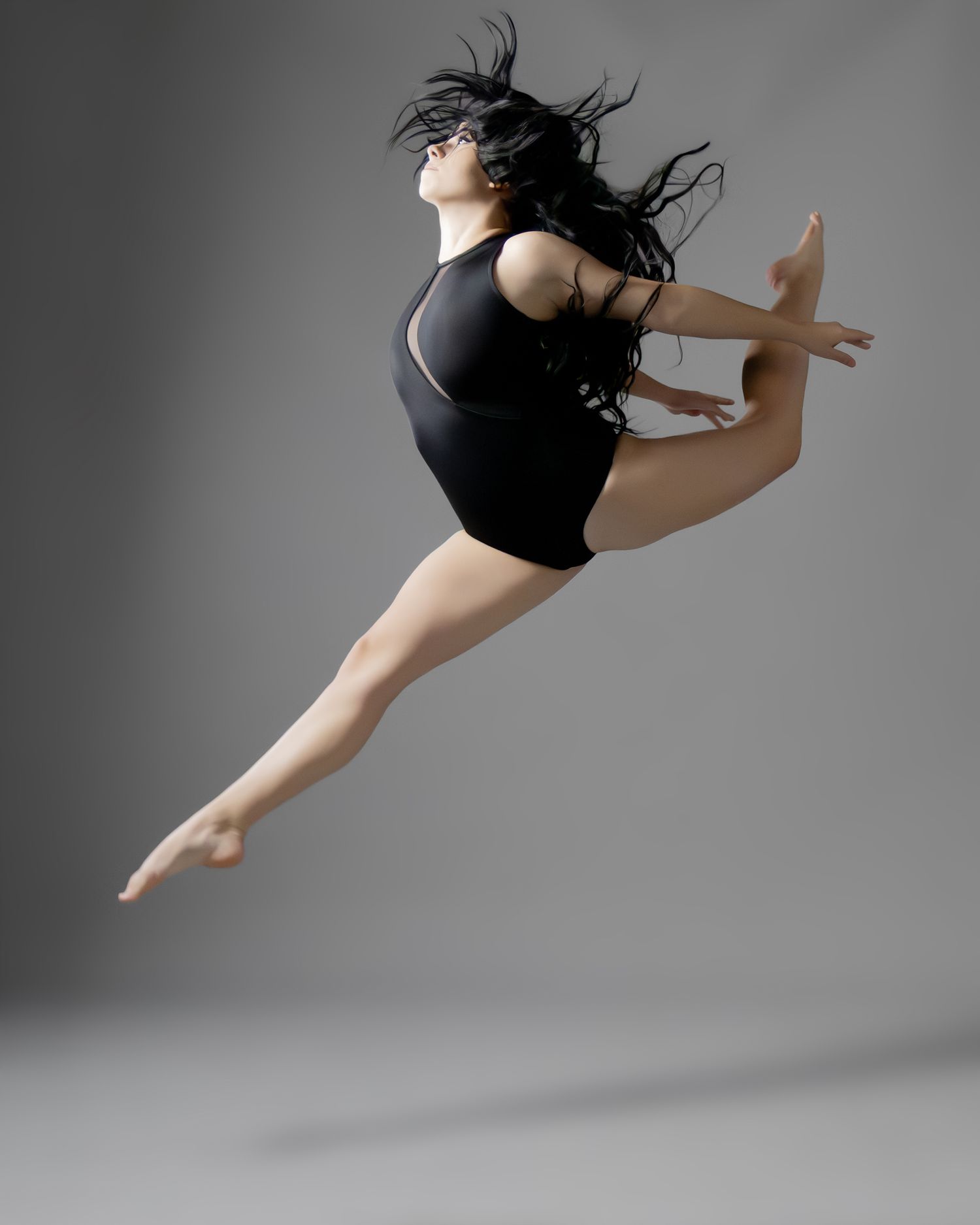 A dancer leaps gracefully in a black leotard, hair flowing, legs extended mid-air against a gray studio background.