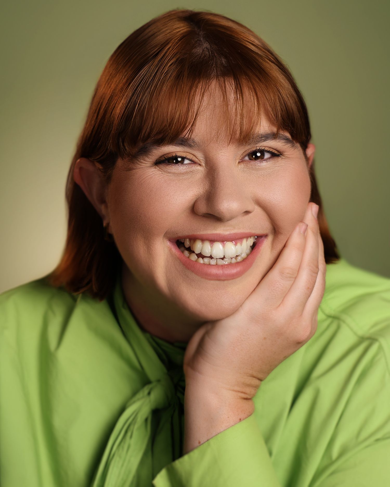 Smiling woman with red hair wearing a bright green top, posing with hand on cheek against green background.
