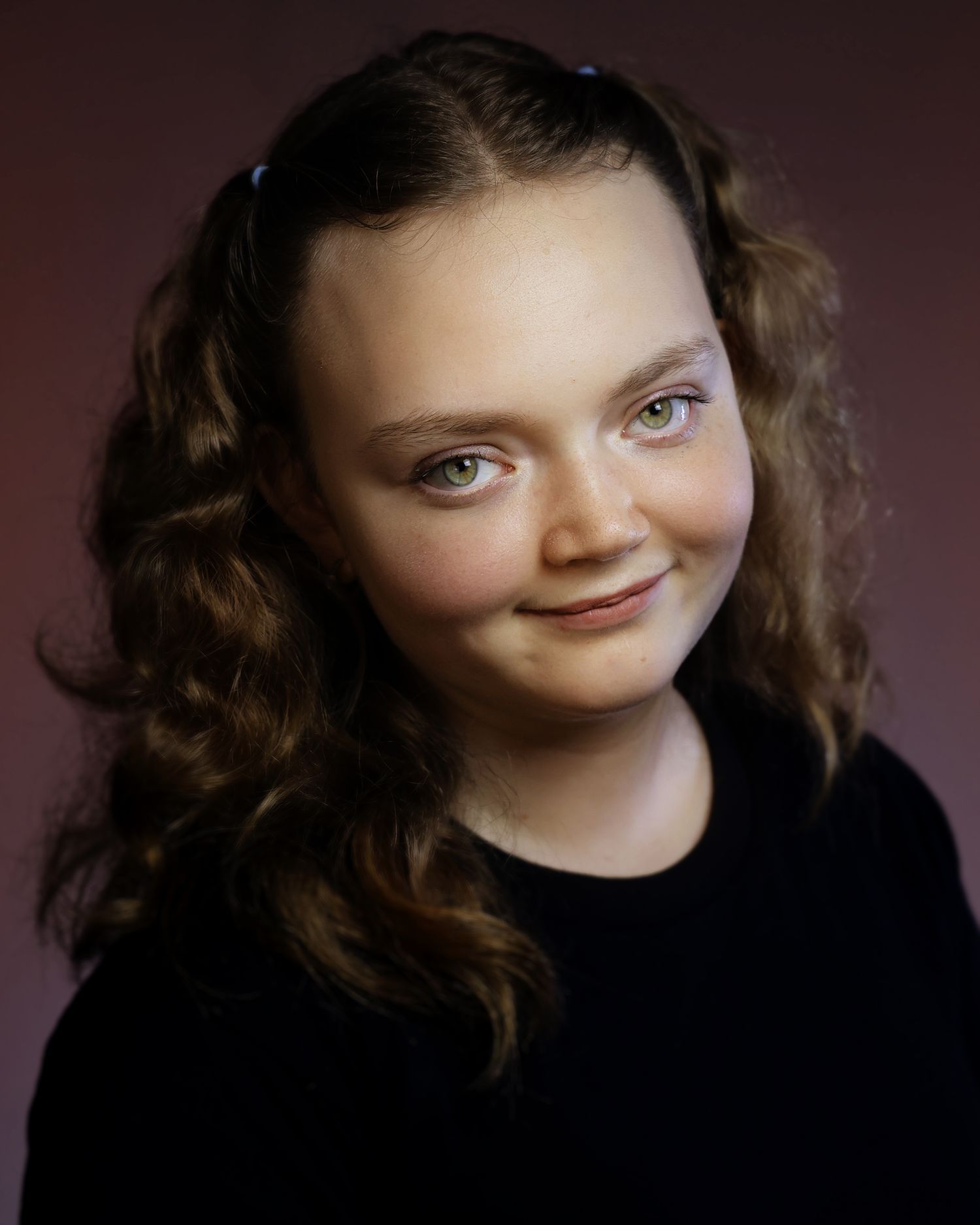 Young girl with curly brown hair and blue clips smiling softly against a purple studio background.