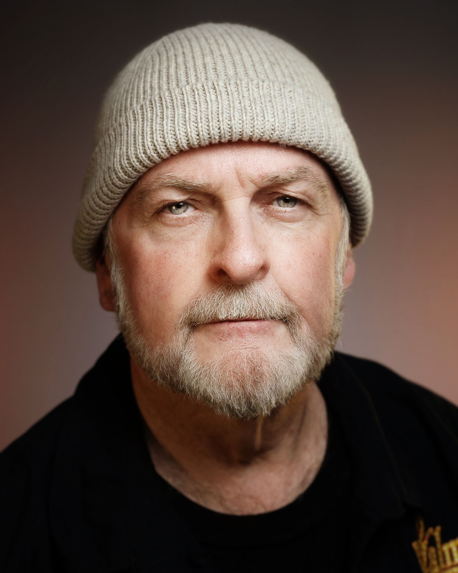 Close-up portrait of a middle-aged man with a gray beard wearing a beige knit beanie hat and black shirt.