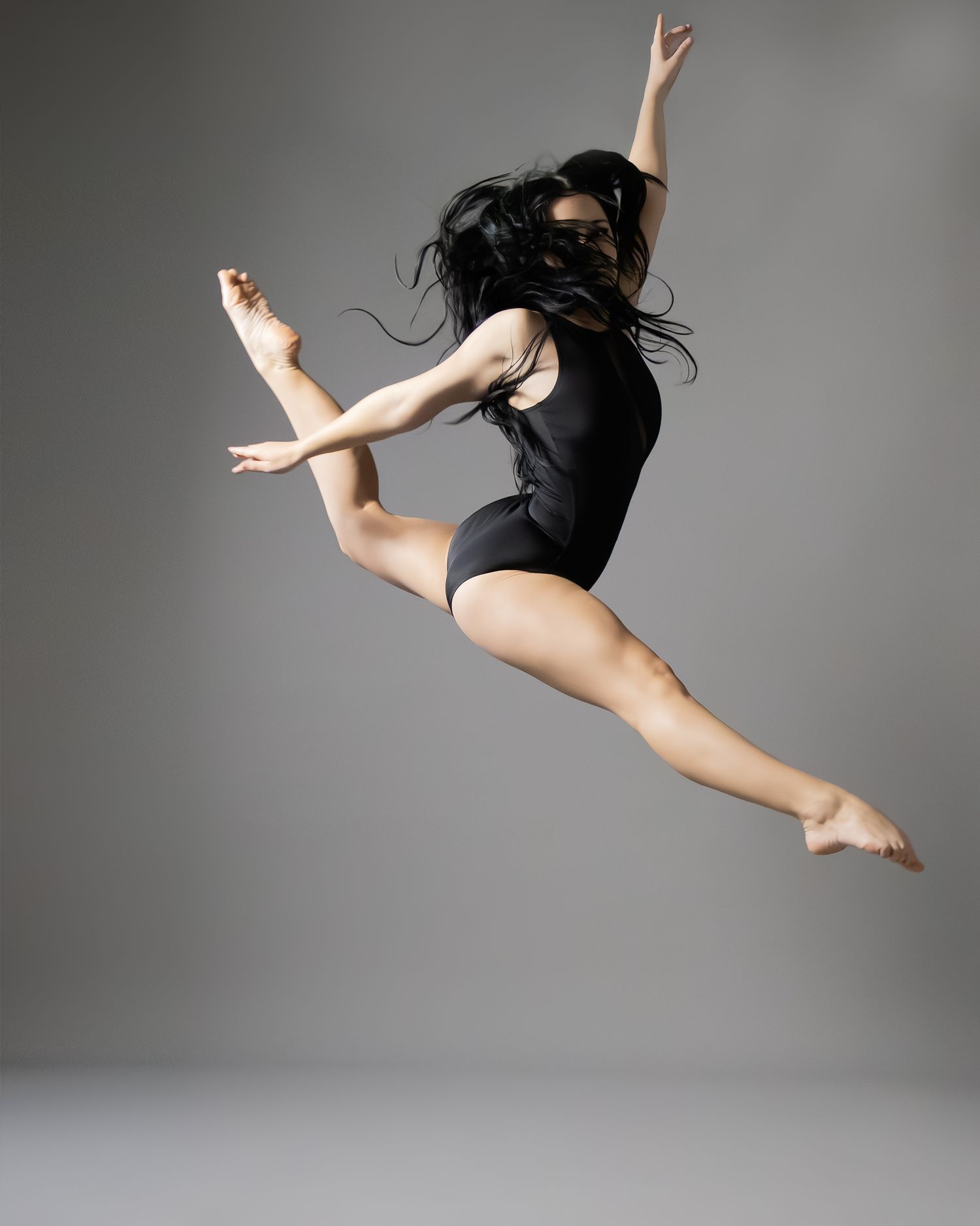A female dancer leaps gracefully in a black leotard against a gray background, hair flowing mid-air.