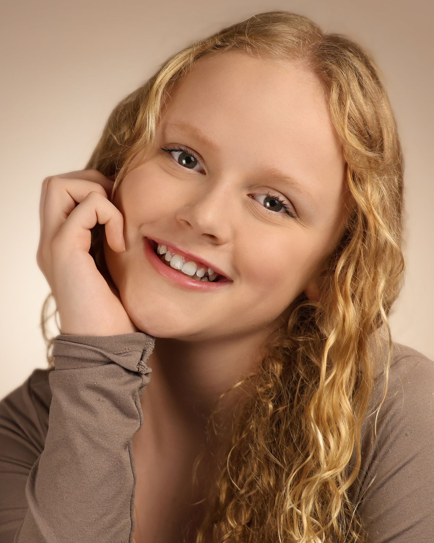 Smiling young girl with curly blonde hair posing with hand on cheek against a neutral background.