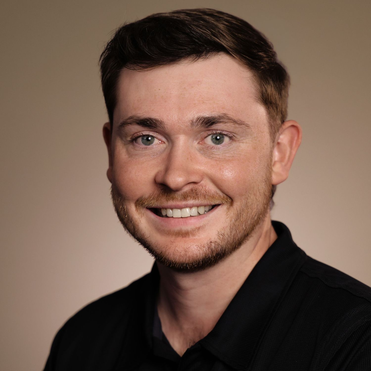 Professional headshot of a smiling young man with short brown hair wearing a black polo shirt against a beige background.