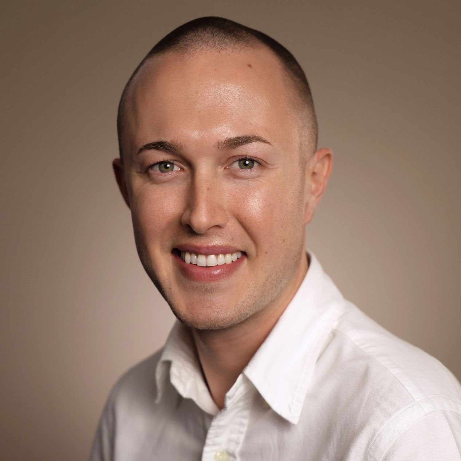 Smiling young man with short hair wearing a white shirt against a neutral beige background.