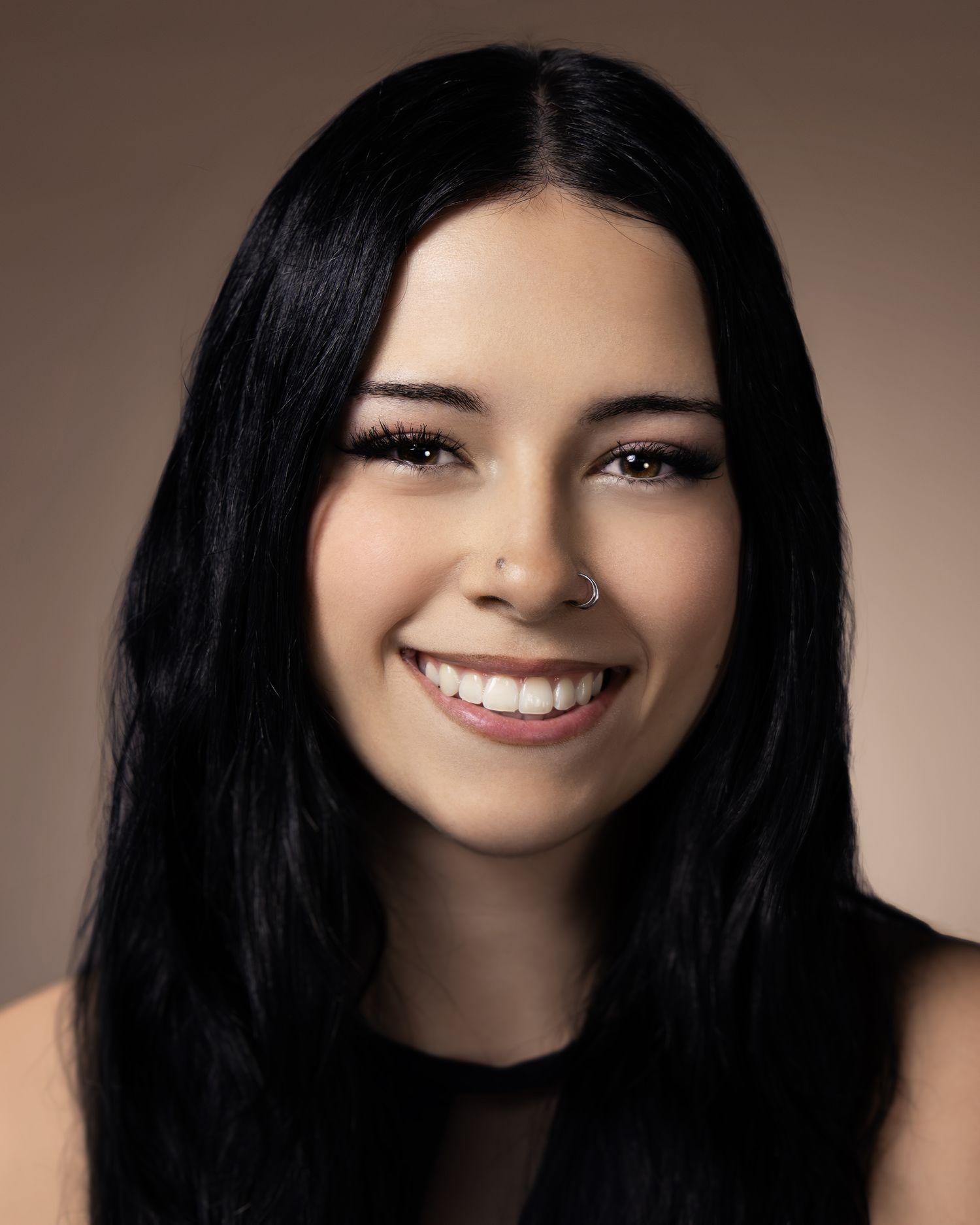 Young woman with long black hair and nose ring smiling confidently against a warm beige background.