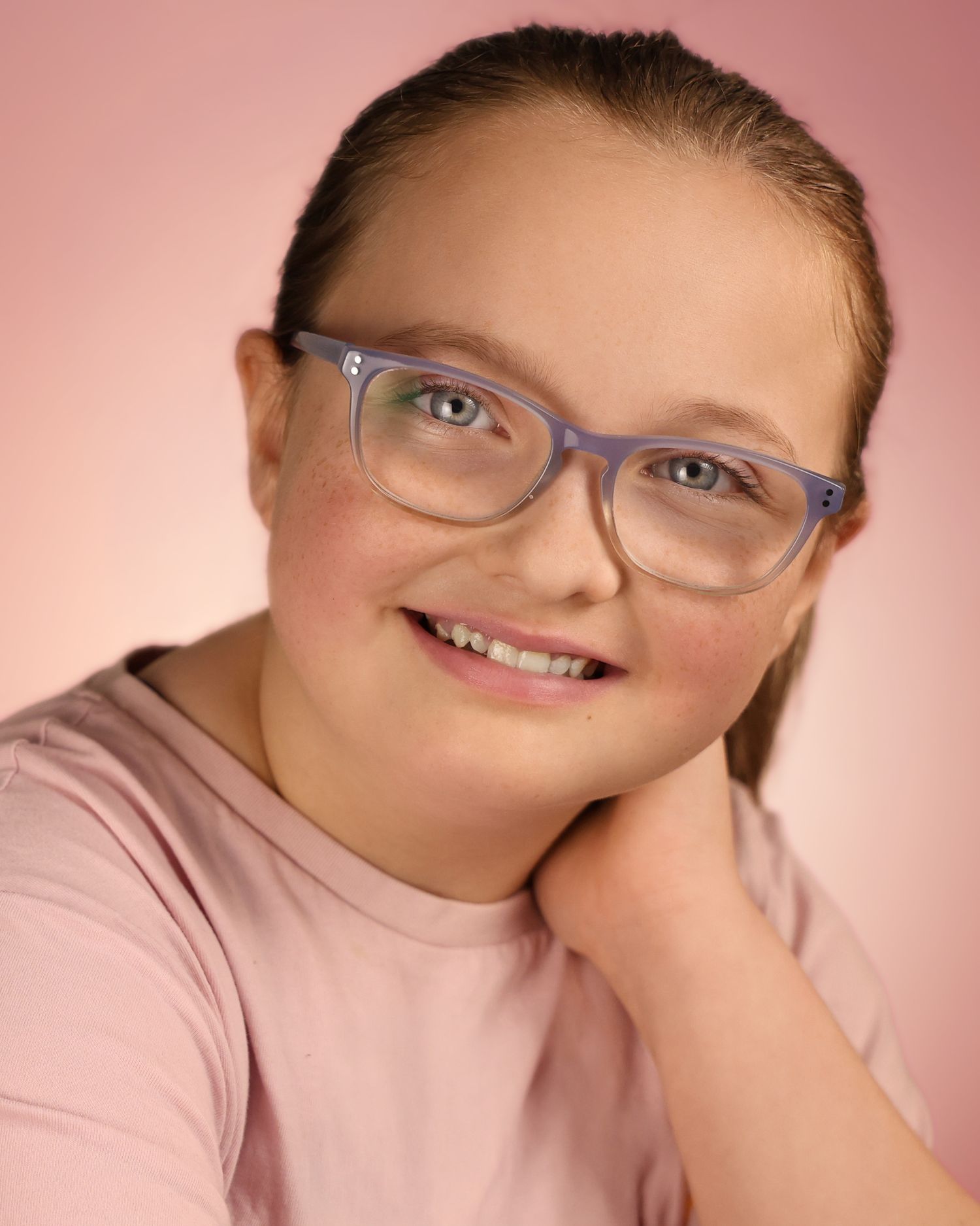 Smiling young girl with Down syndrome wearing purple glasses and pink shirt poses for a portrait.