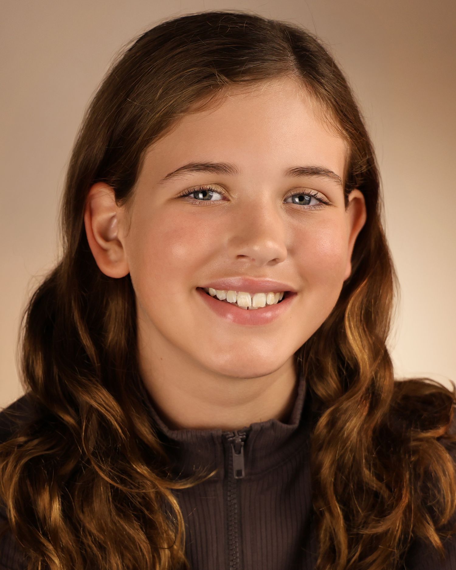Smiling young girl with long curly brown hair wearing a dark zip-up jacket, posed for a portrait photo.