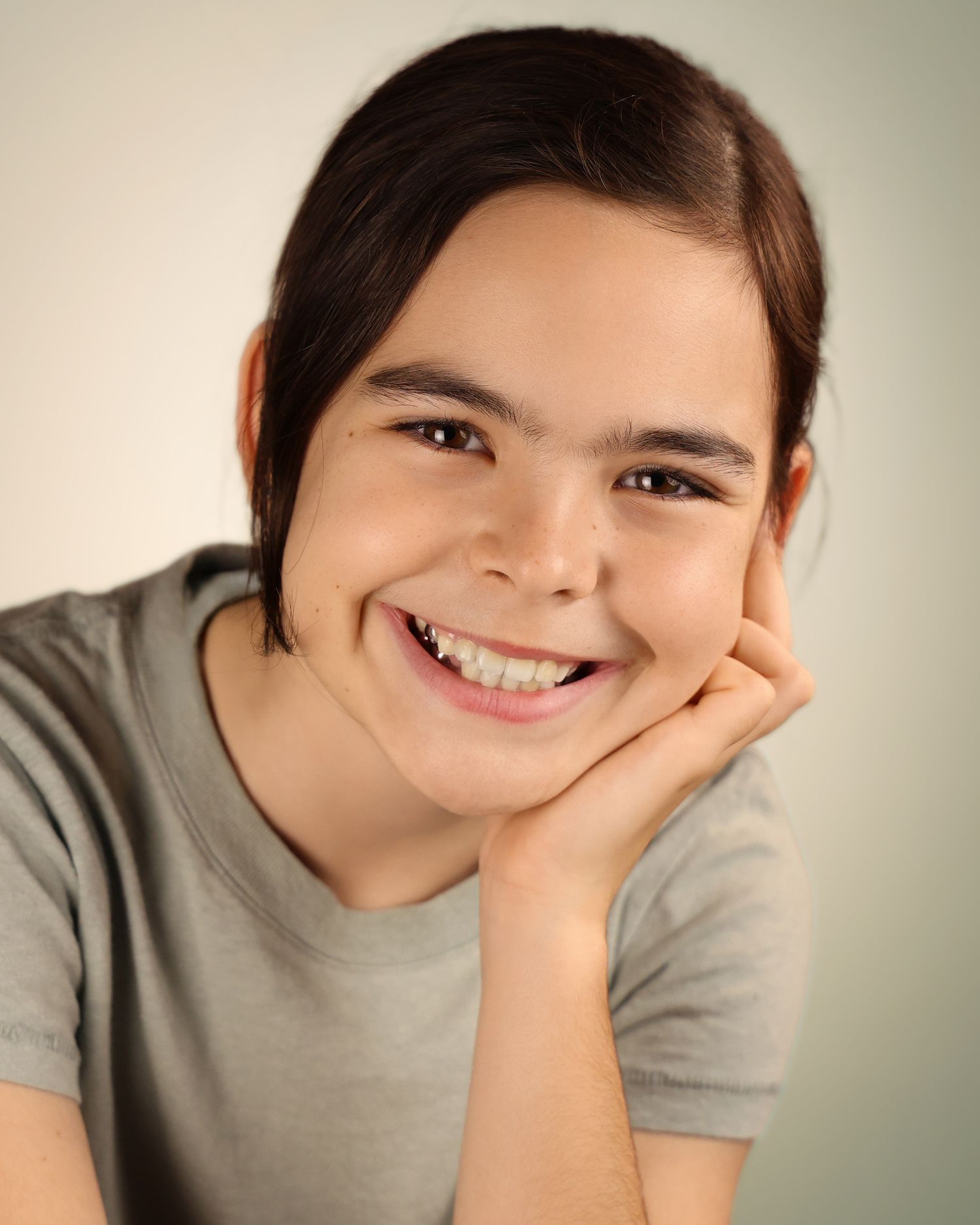 Smiling young girl with dark hair resting chin on hand, wearing a gray t-shirt, studio portrait photo.